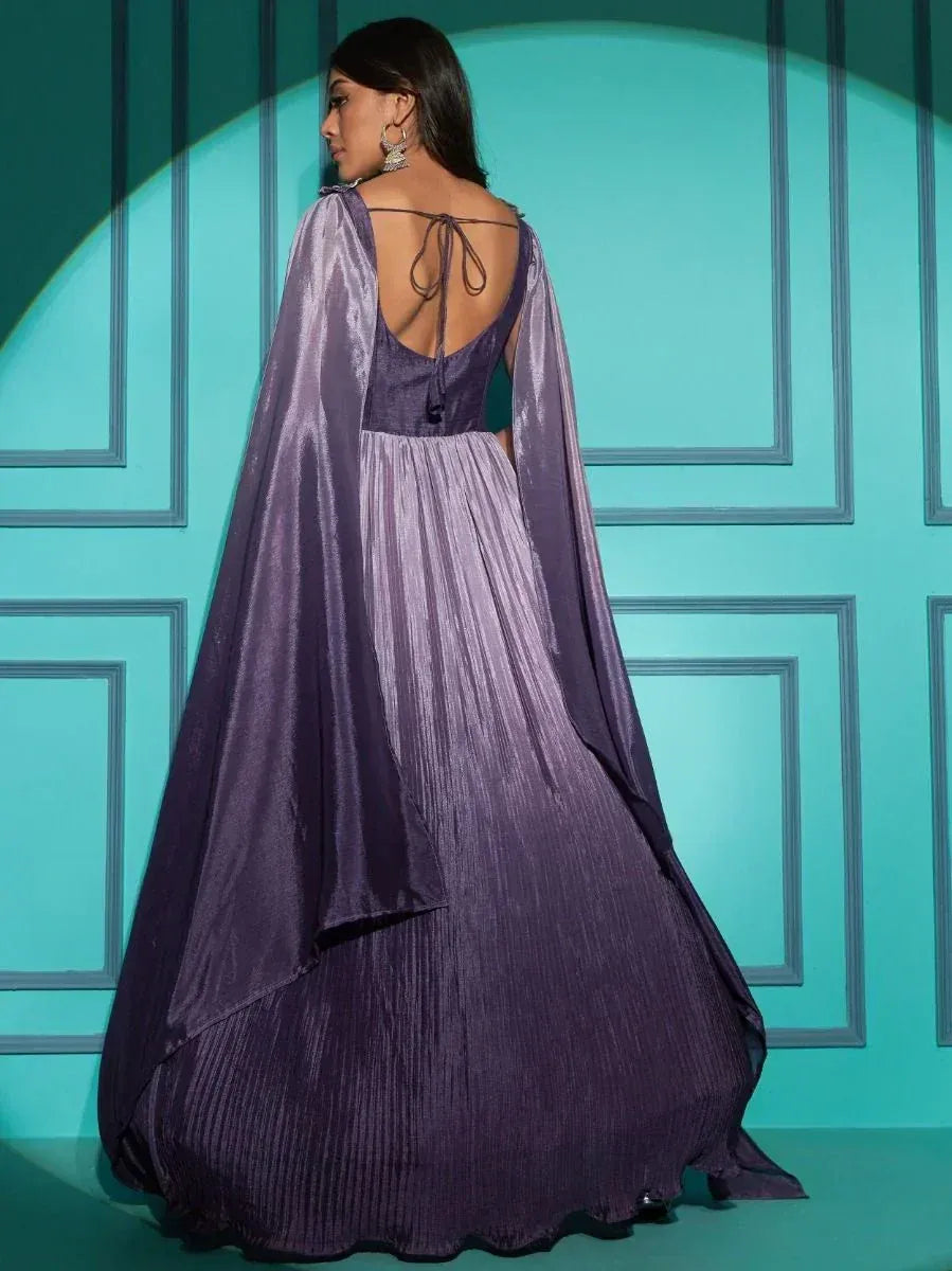 Woman in a purple pleated ethnic gown with cape sleeves, open tie-back, standing against a teal wall