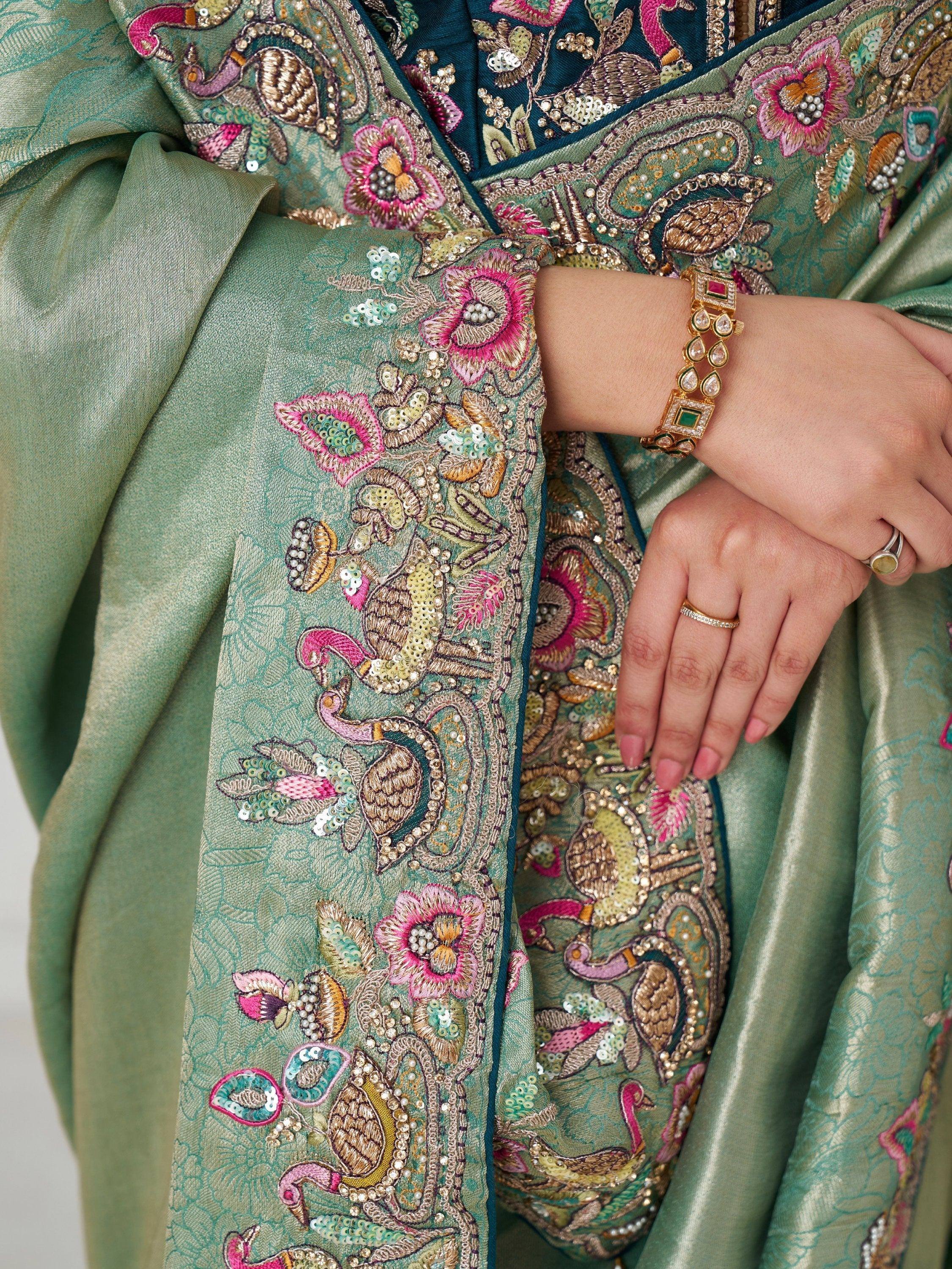 Green embroidered saree with peacock and floral motifs, sequins, and pearl detailing, paired with gold jewelry.