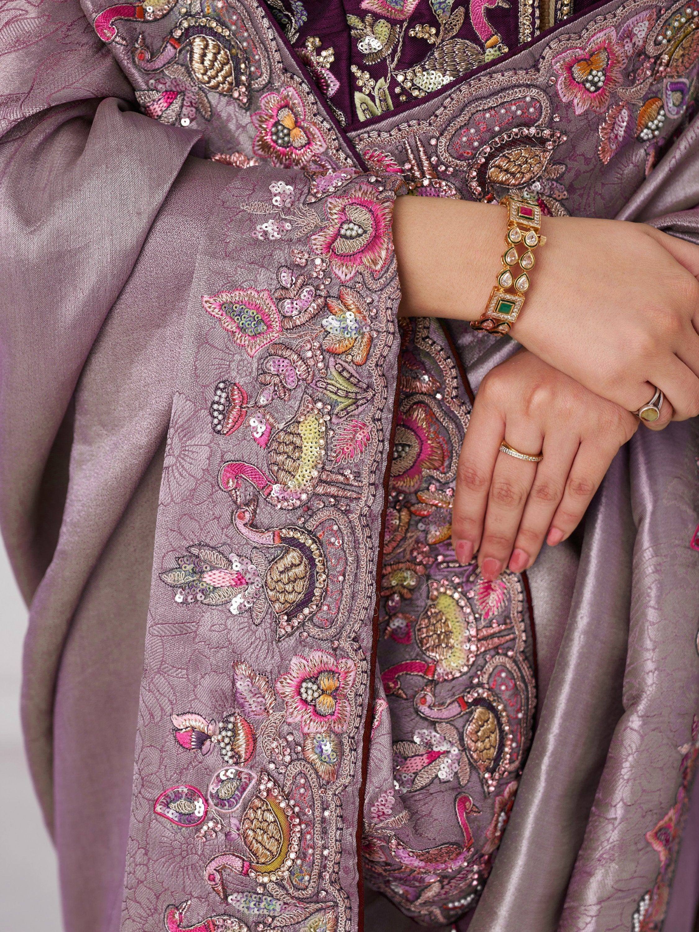 Purple ethnic gown with peacock embroidery, sequins, and floral motifs, paired with gold jewelry.