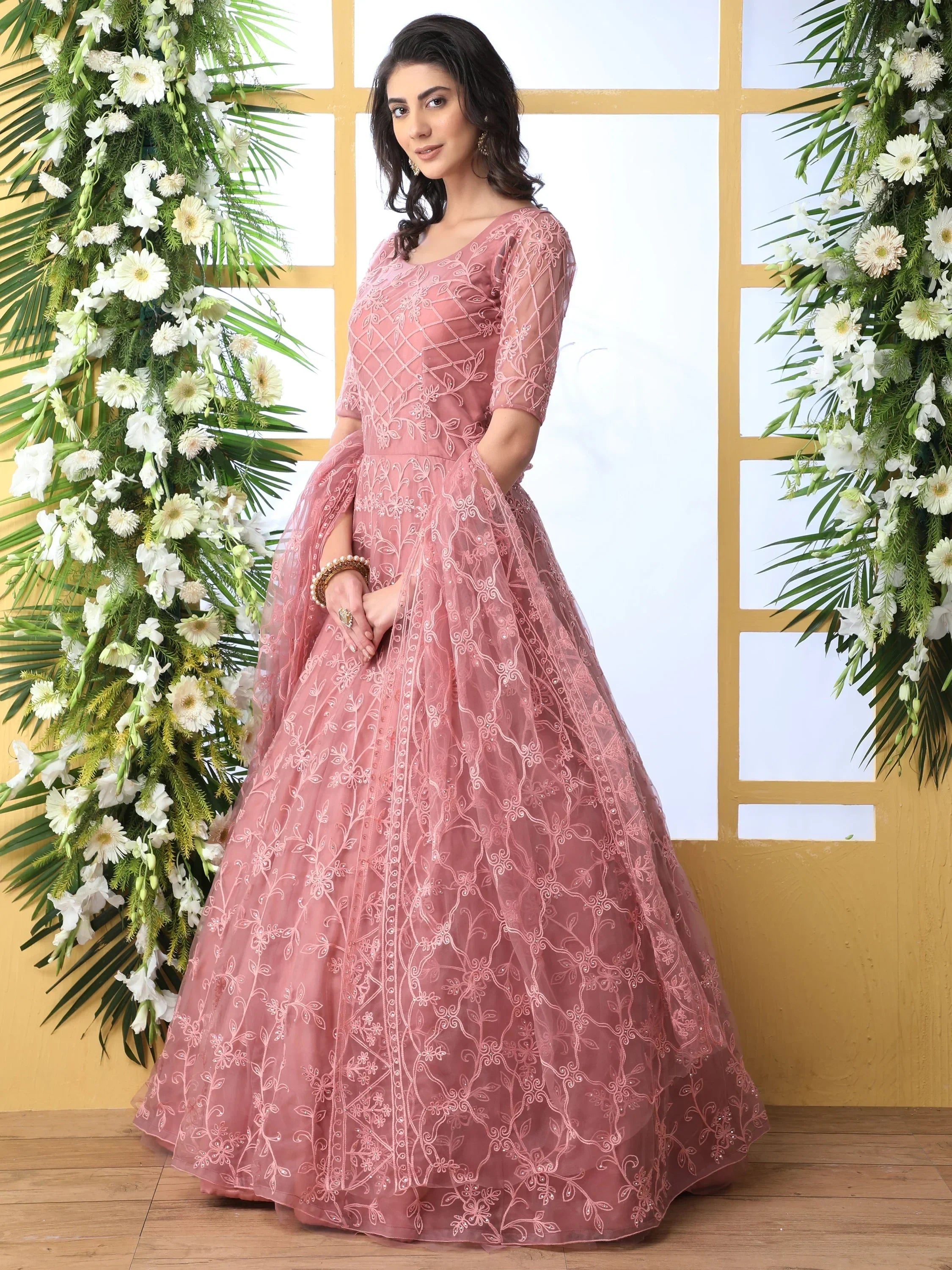 Woman wearing pink embroidered lehenga gown with floral patterns, standing by flower decor