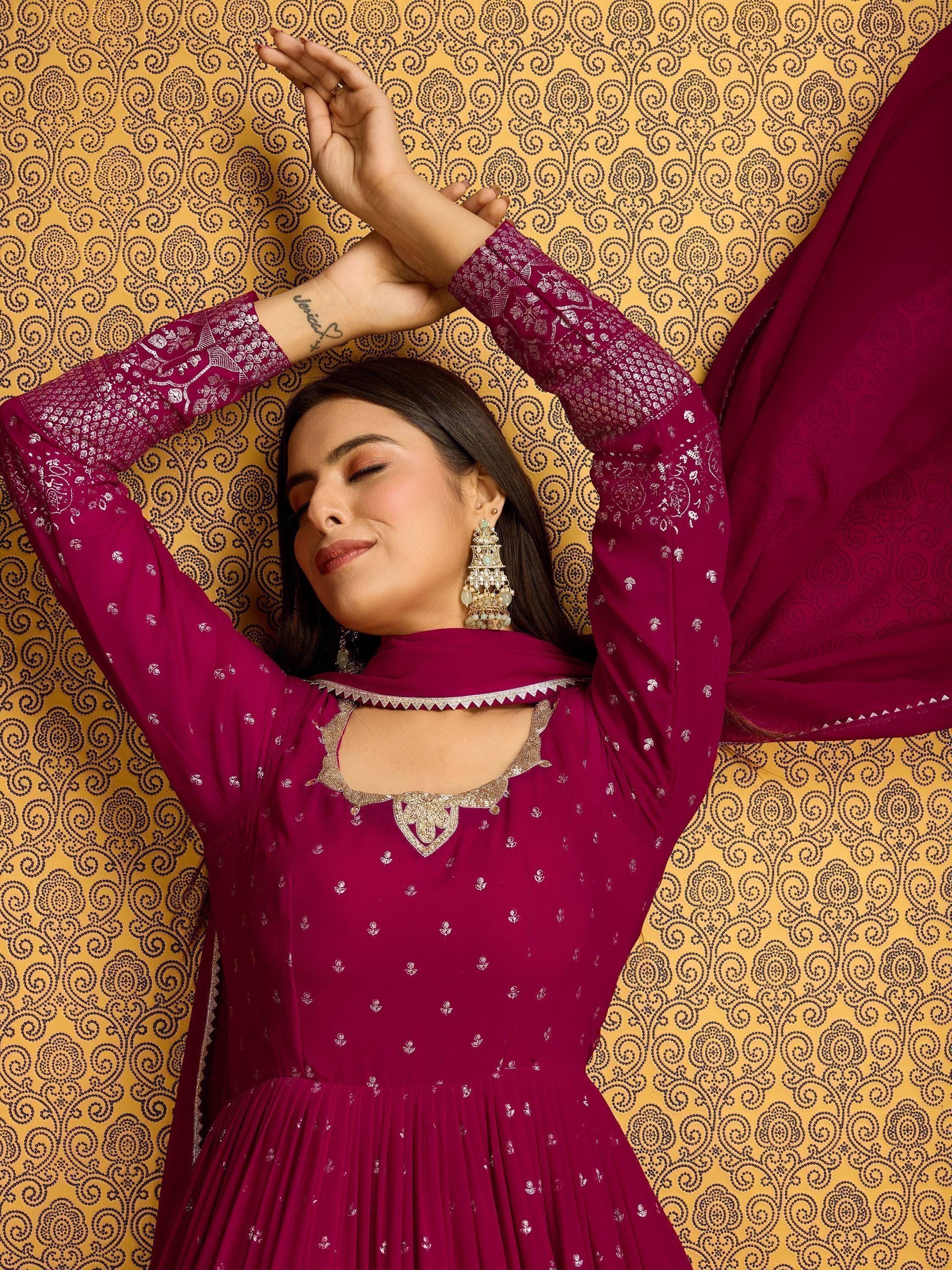 Woman in maroon ethnic gown with silver embroidery, paired with statement earrings, against patterned backdrop