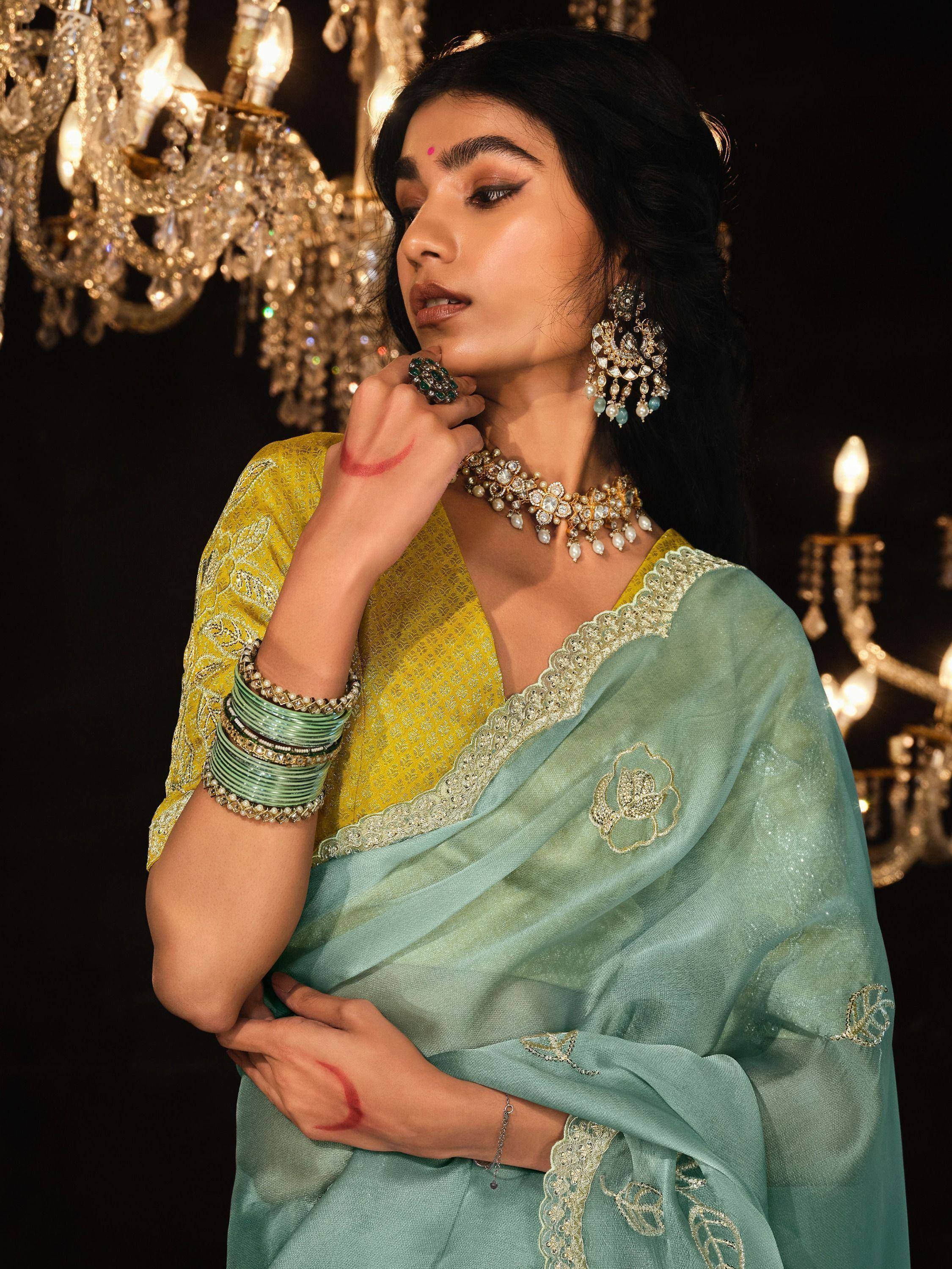 Elegant woman in mint green saree with gold embroidery, paired with yellow blouse and traditional jewelry, under chandelier