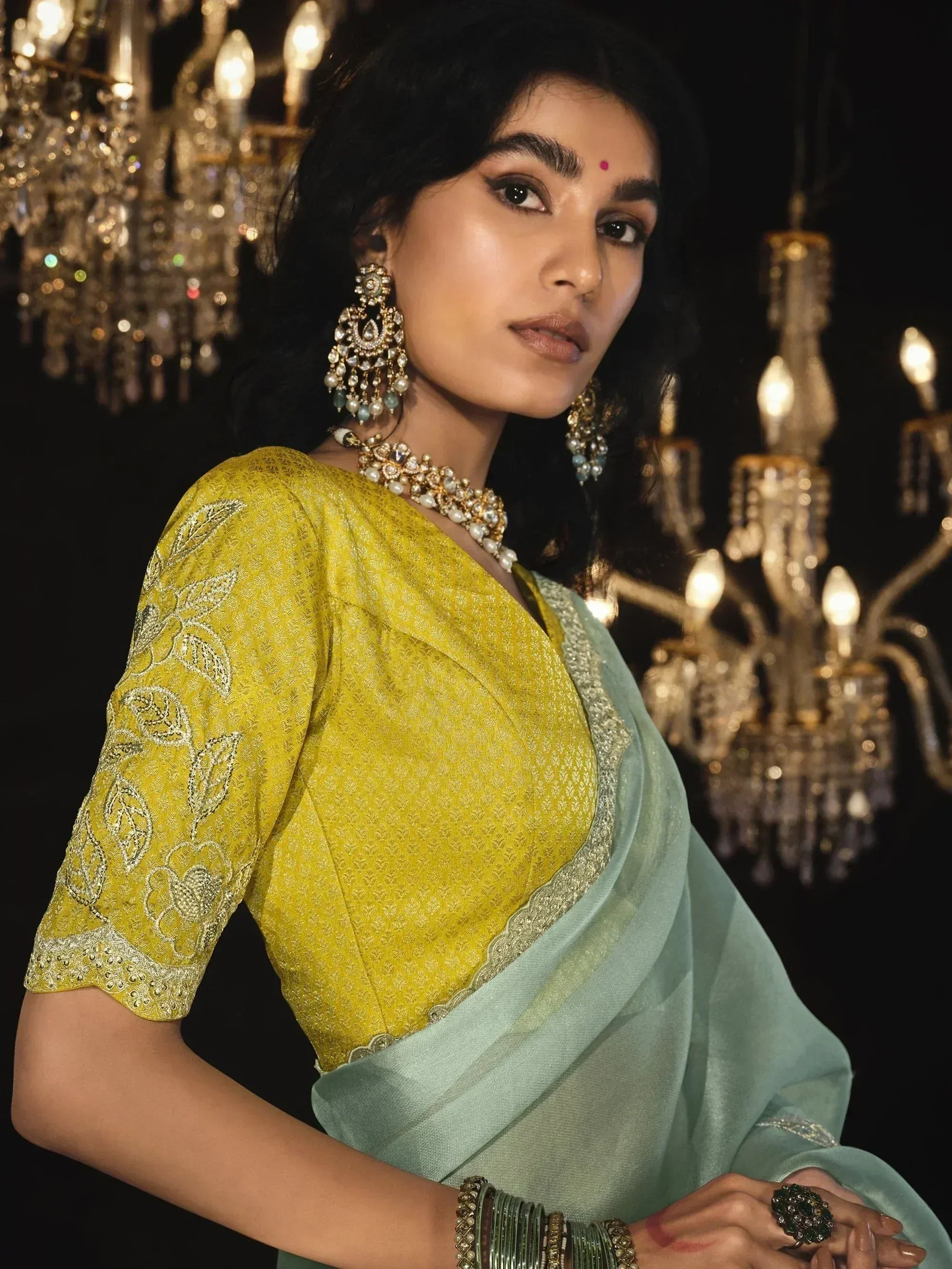 Elegant woman in yellow embroidered blouse and mint saree with traditional jewelry under chandelier