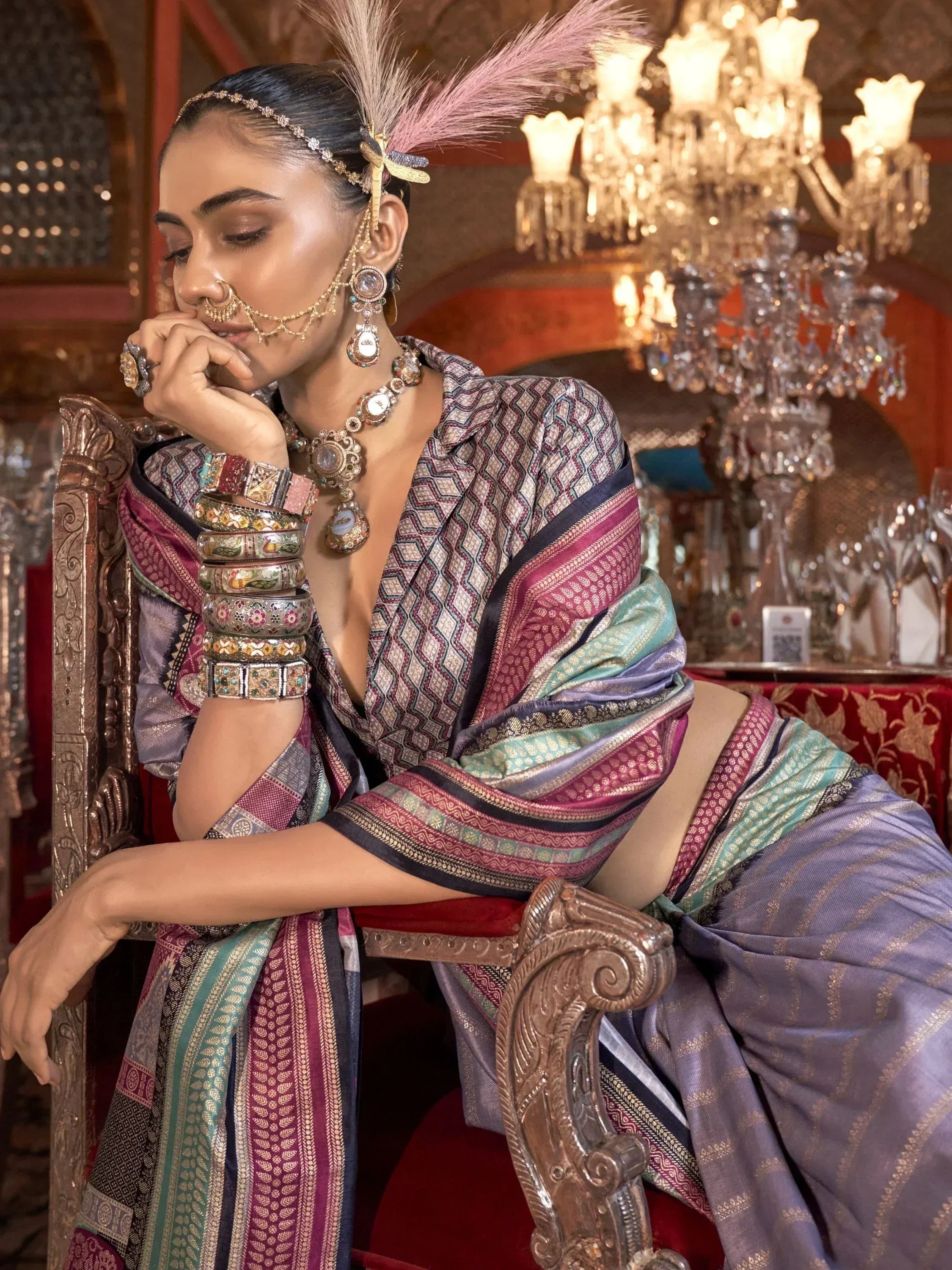 Elegant woman in colorful ethnic saree with intricate jewelry and feather headpiece, seated on ornate chair.