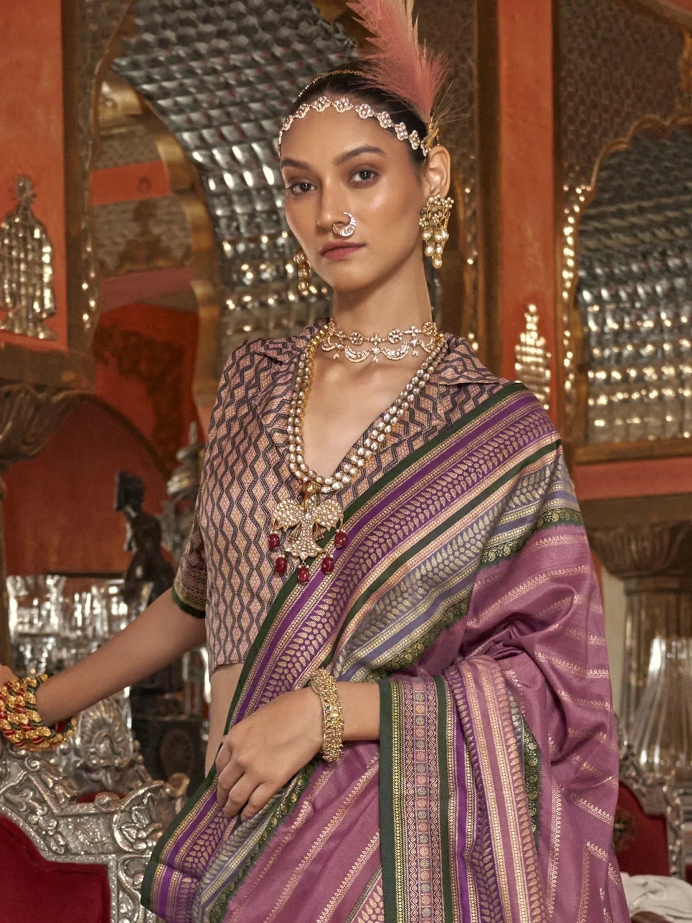 Elegant woman in purple and green patterned saree with gold jewelry and feather headpiece, showcasing Indian ethnic wear.