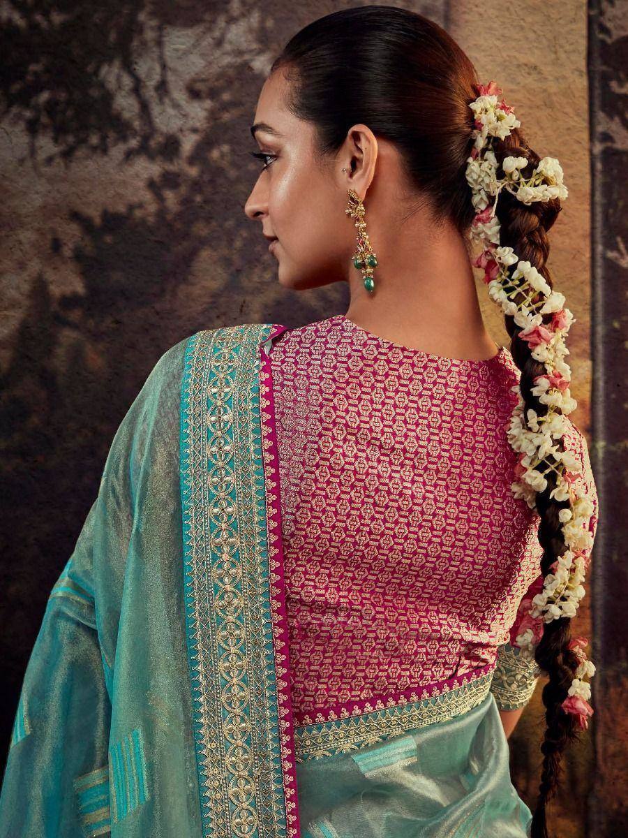 Woman in pink patterned blouse with teal saree, floral braid, and gold earrings