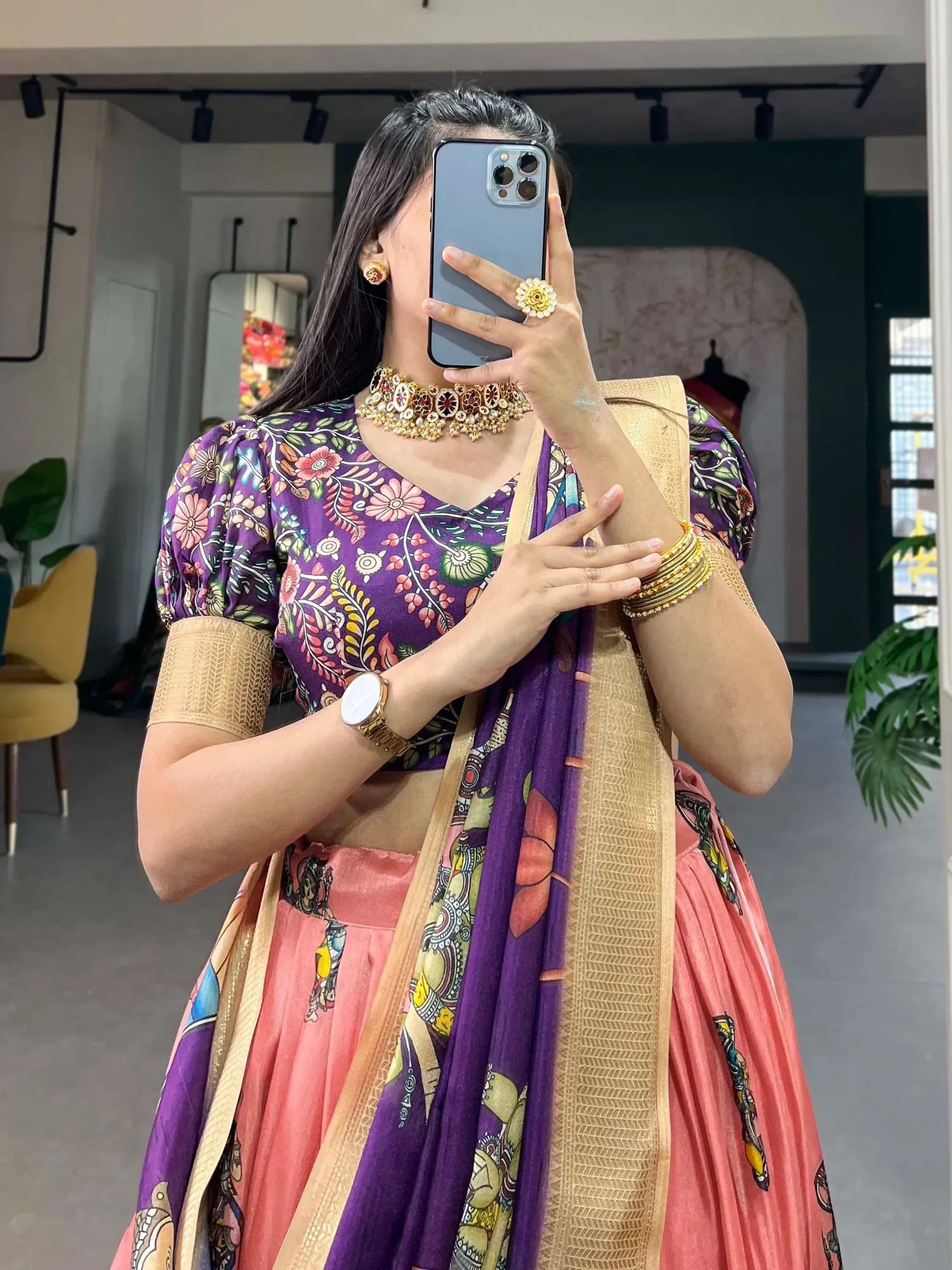 Woman in purple floral blouse and pink lehenga with gold dupatta, taking mirror selfie in ethnic wear store