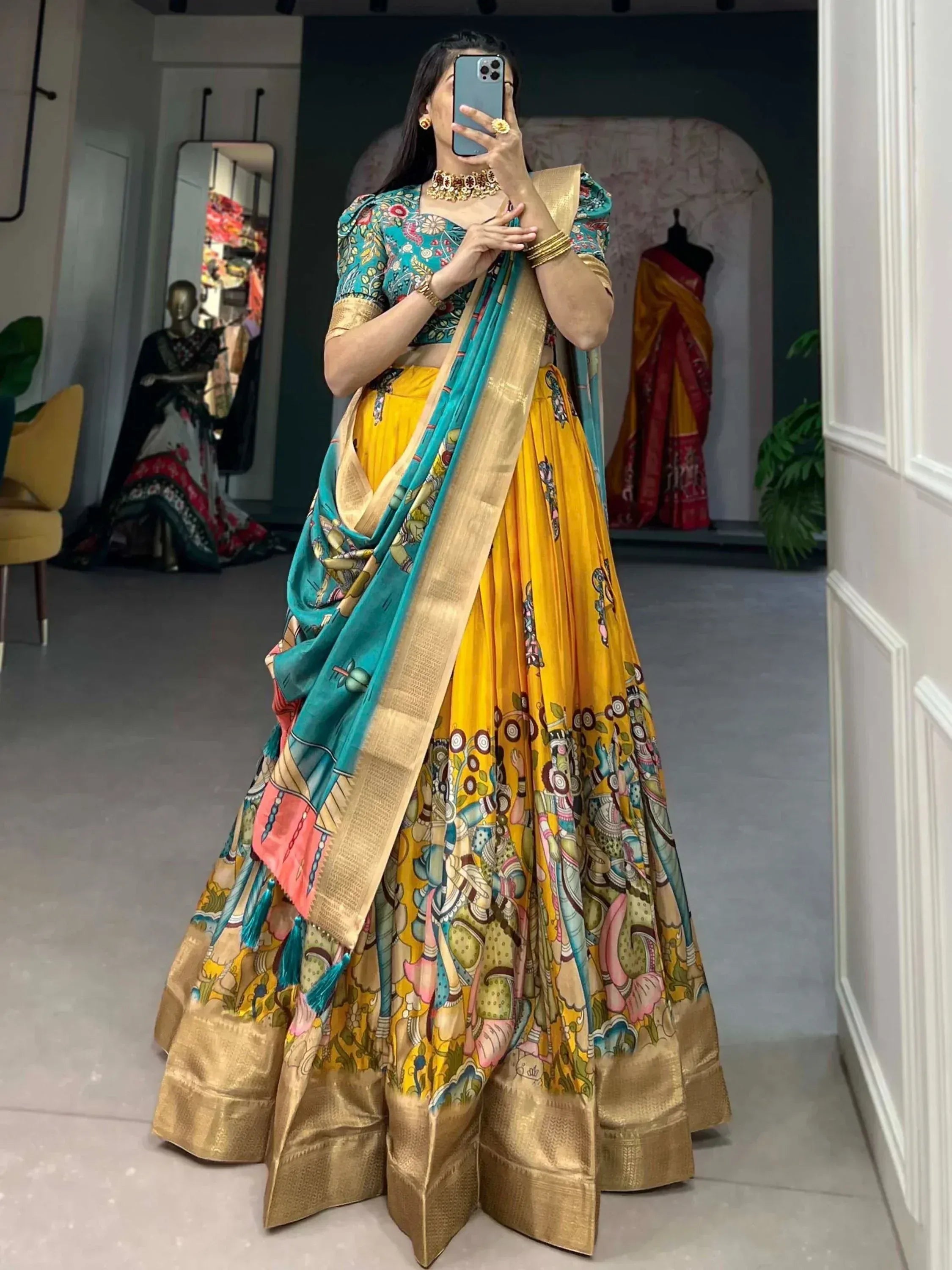 Woman in vibrant yellow and teal printed lehenga choli with gold border, taking mirror selfie in boutique