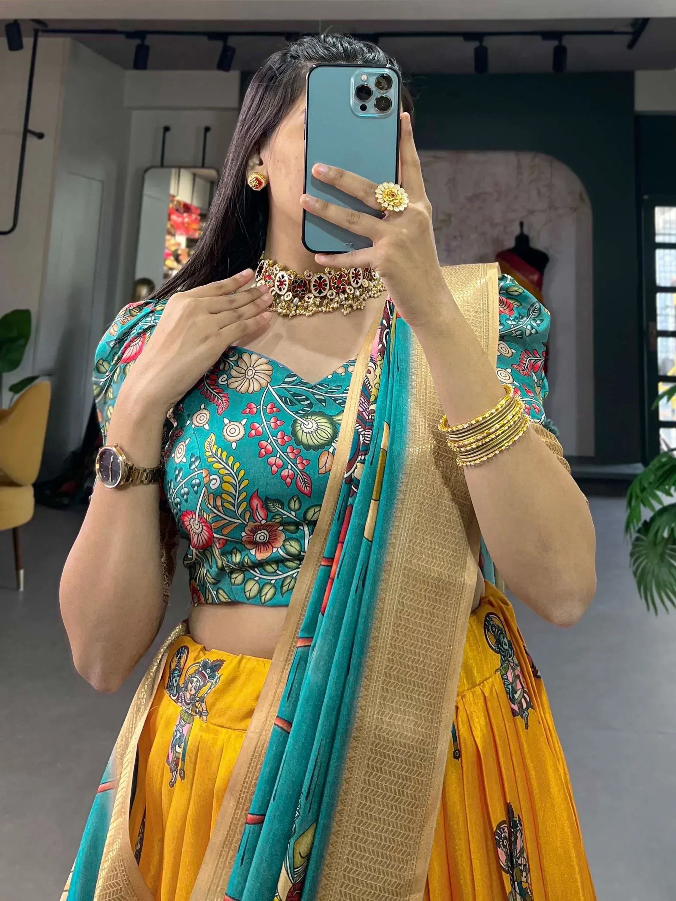 Woman in vibrant teal and yellow printed lehenga choli with gold jewelry, taking mirror selfie