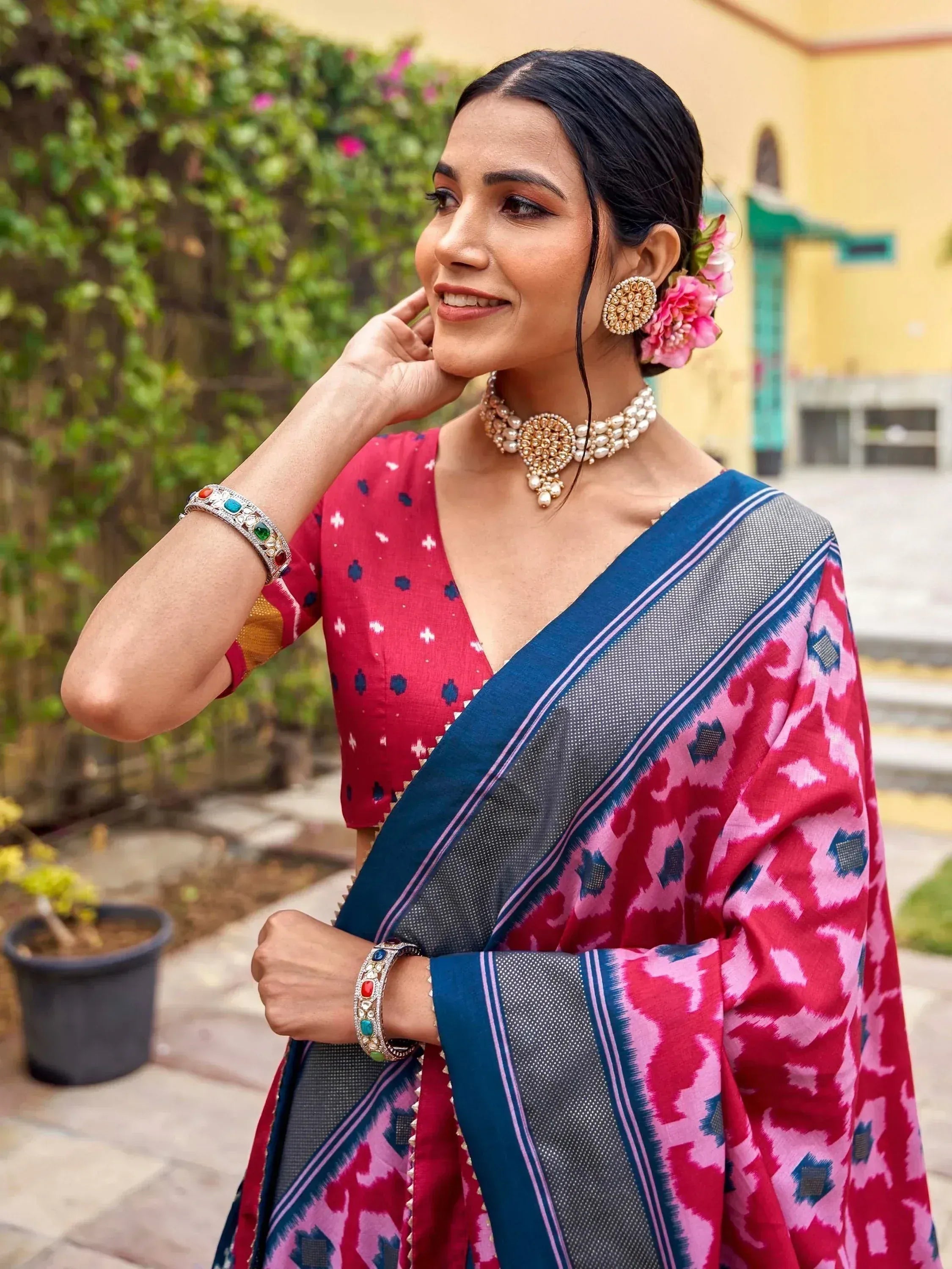 Woman in vibrant pink and blue ikat saree with pearl jewelry, floral hair accessory, and traditional bangles.