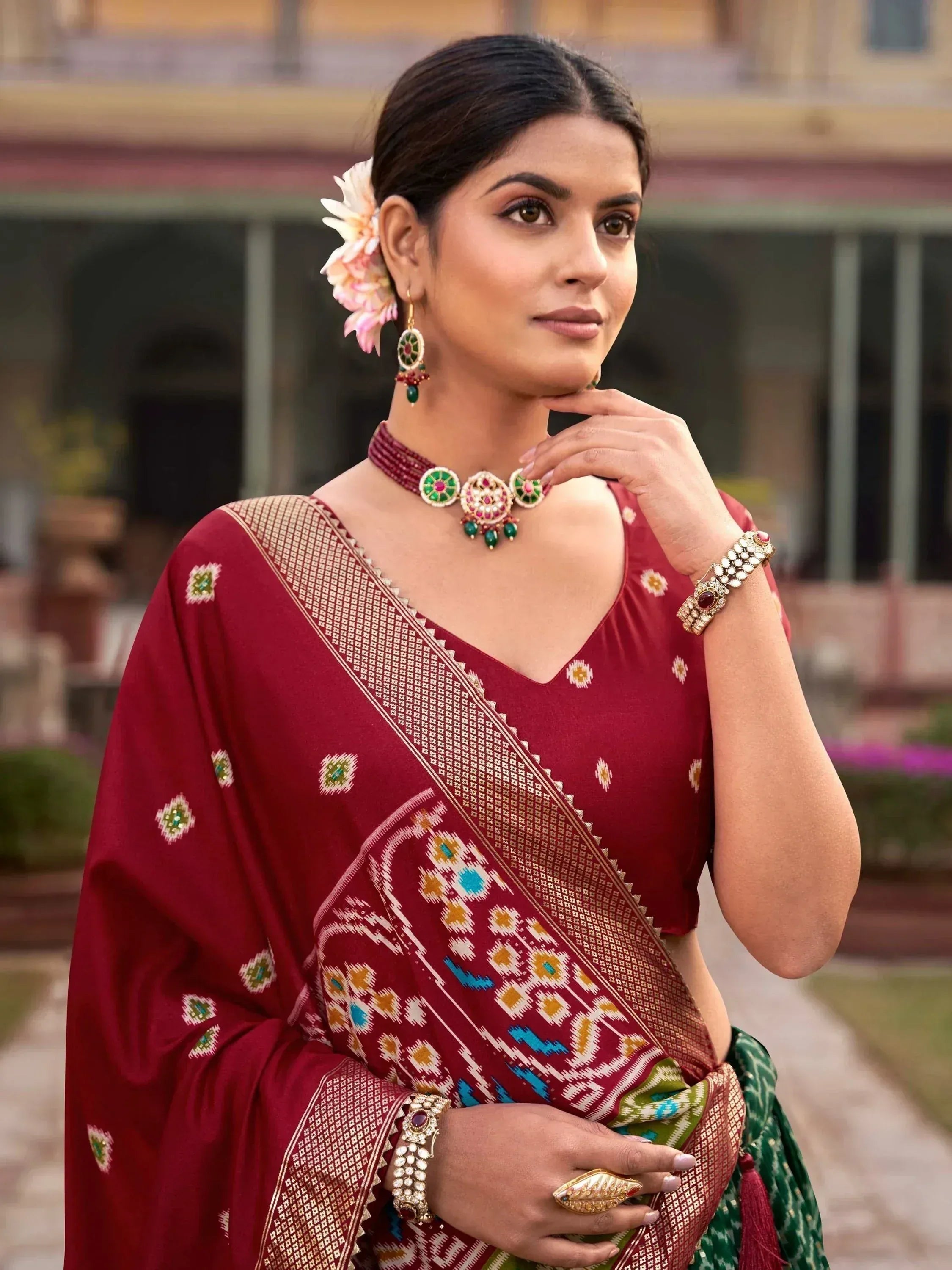 Woman in maroon ethnic saree with gold and green embroidery, traditional jewelry, and floral hair accessory
