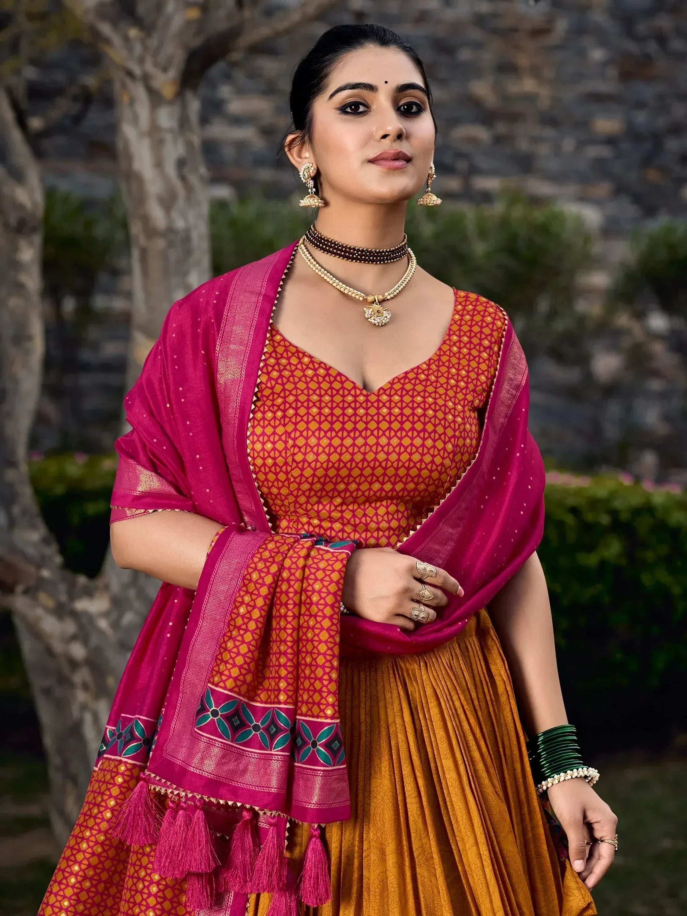 Woman in vibrant Indian ethnic lehenga choli with pink dupatta, traditional jewelry, and tassels, perfect for festive occasions.