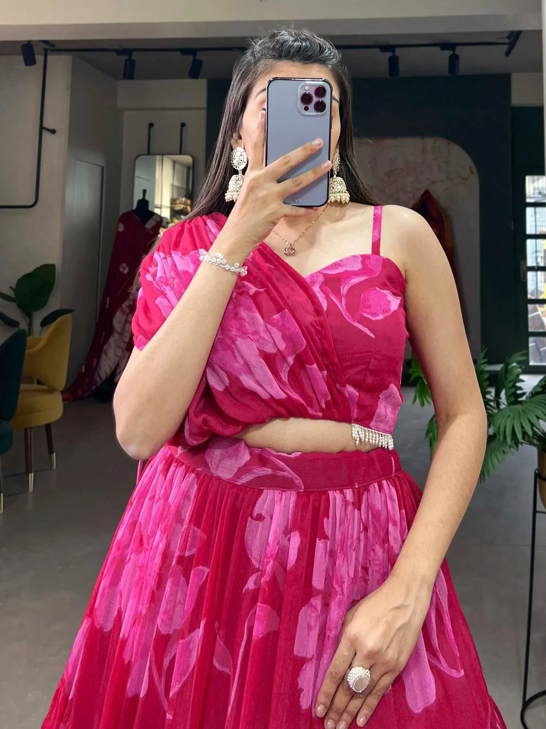 Woman in vibrant pink floral lehenga choli with pearl jewelry, taking mirror selfie in boutique