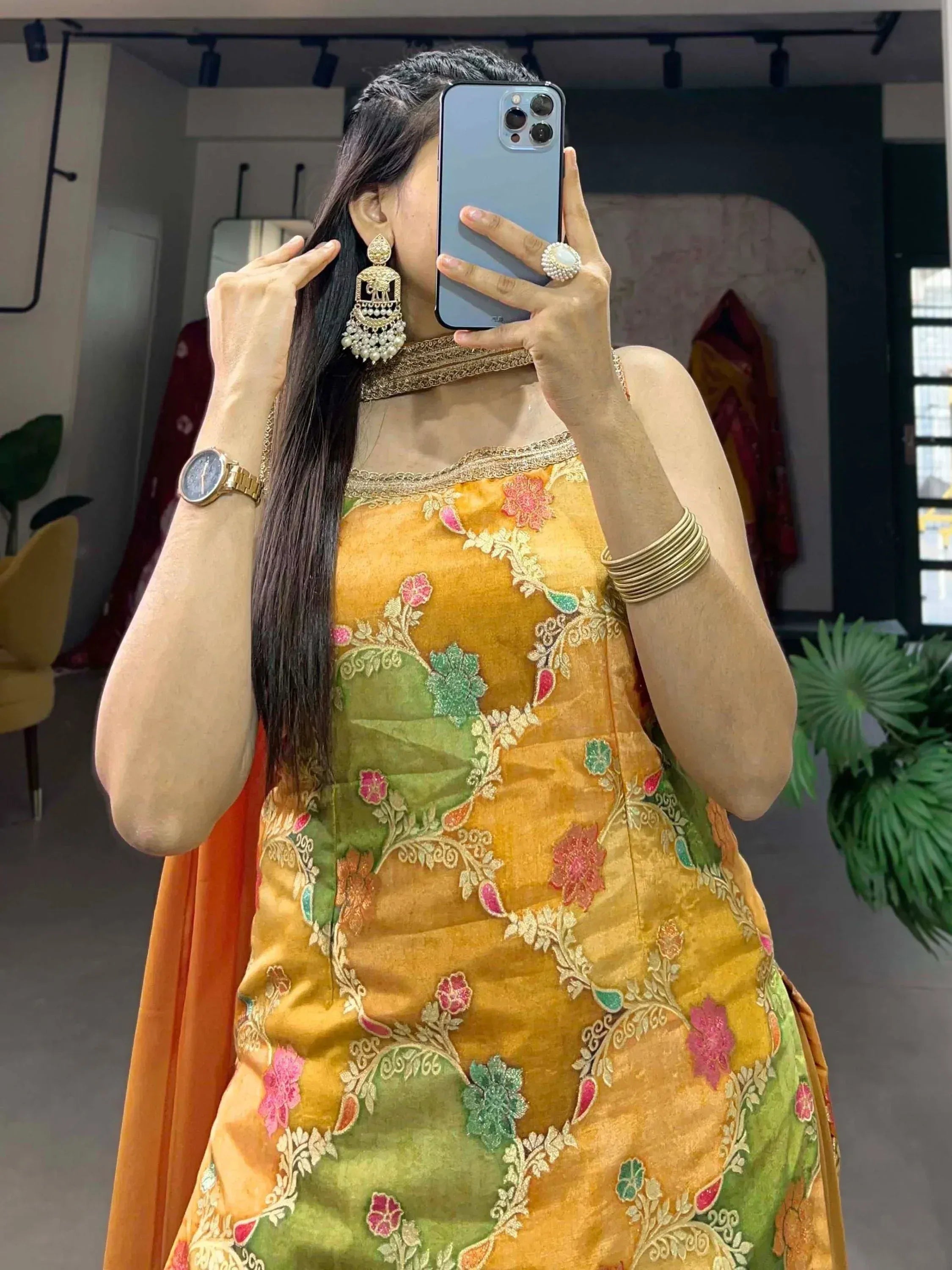 Woman in mustard yellow floral embroidered ethnic gown with gold jewelry, taking mirror selfie