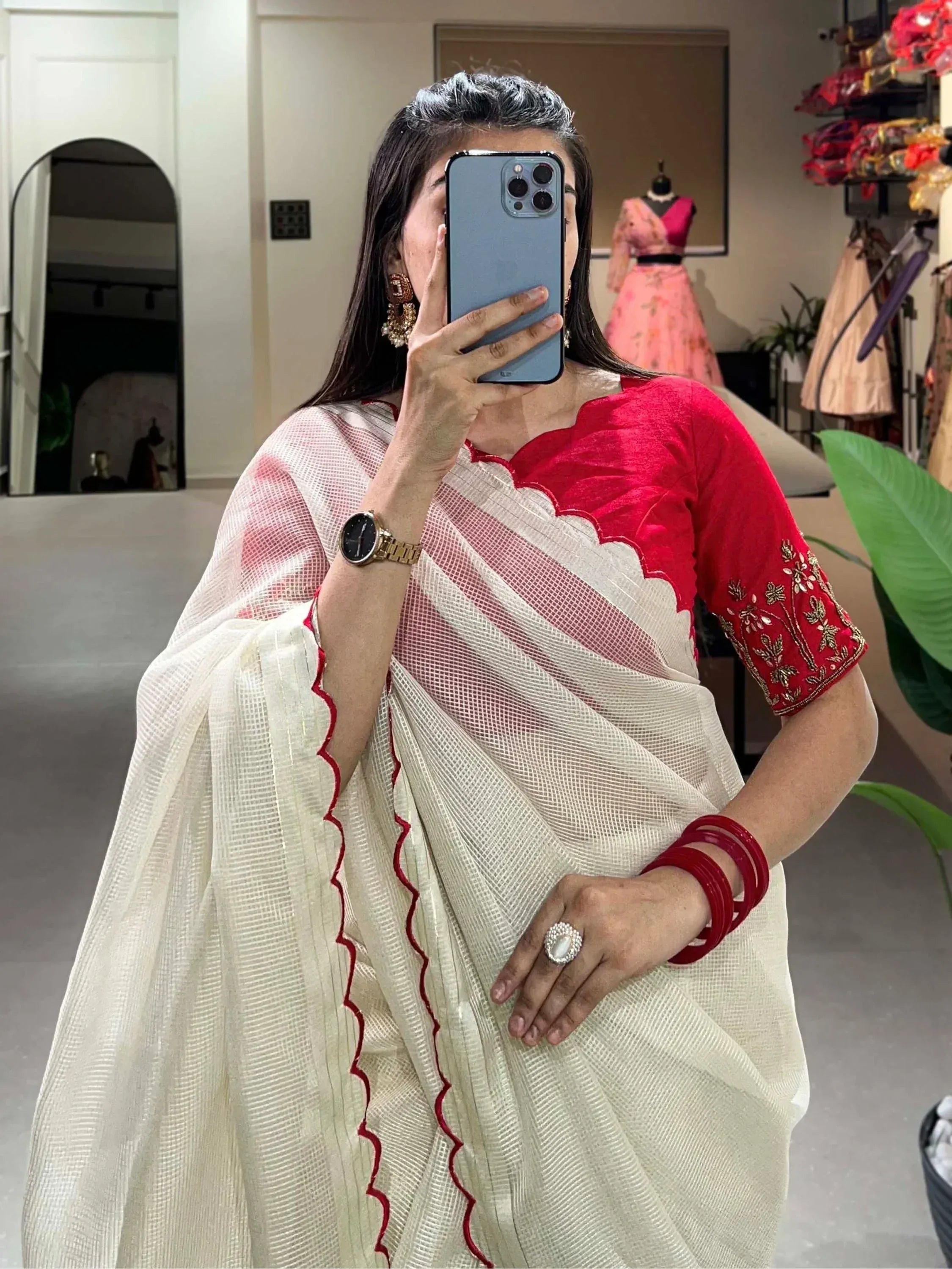 Woman in red embroidered blouse and white saree with red trim, taking mirror selfie in boutique