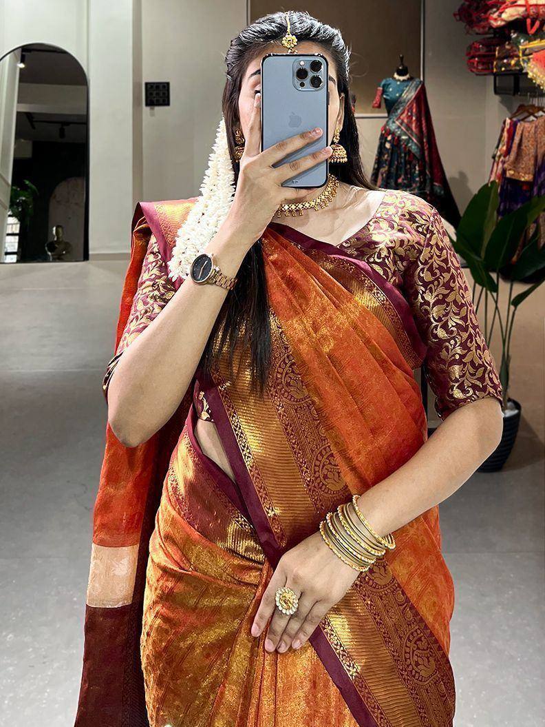 Woman in orange and maroon designer saree with gold embroidery, traditional jewelry, and floral garland