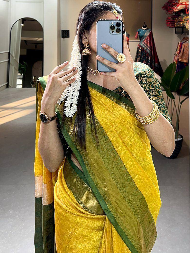 Woman in yellow and green ethnic saree with floral hair accessory, taking mirror selfie in boutique