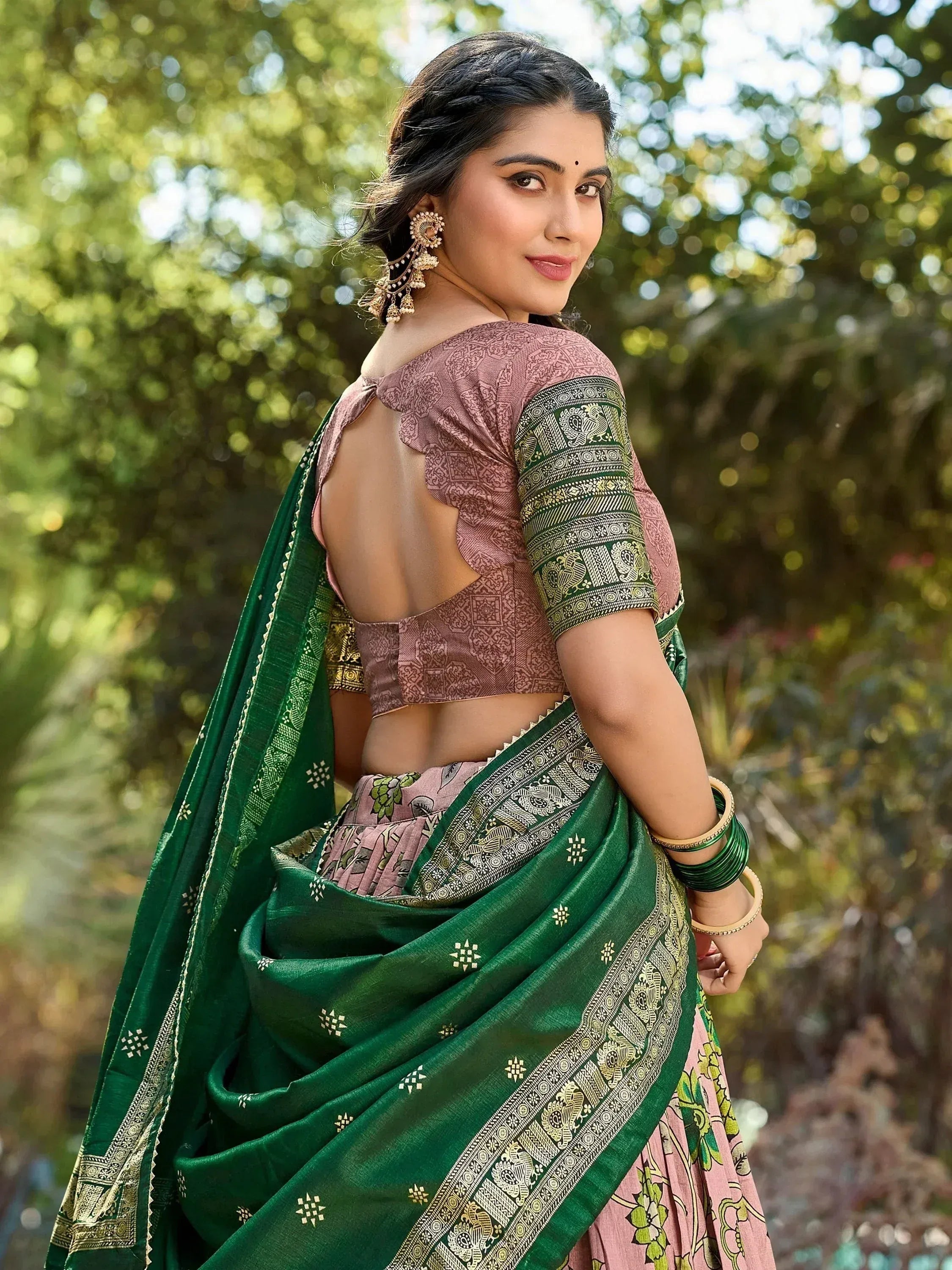 Woman in green and pink ethnic saree with backless blouse, floral print, and gold border, styled with jhumka earrings.