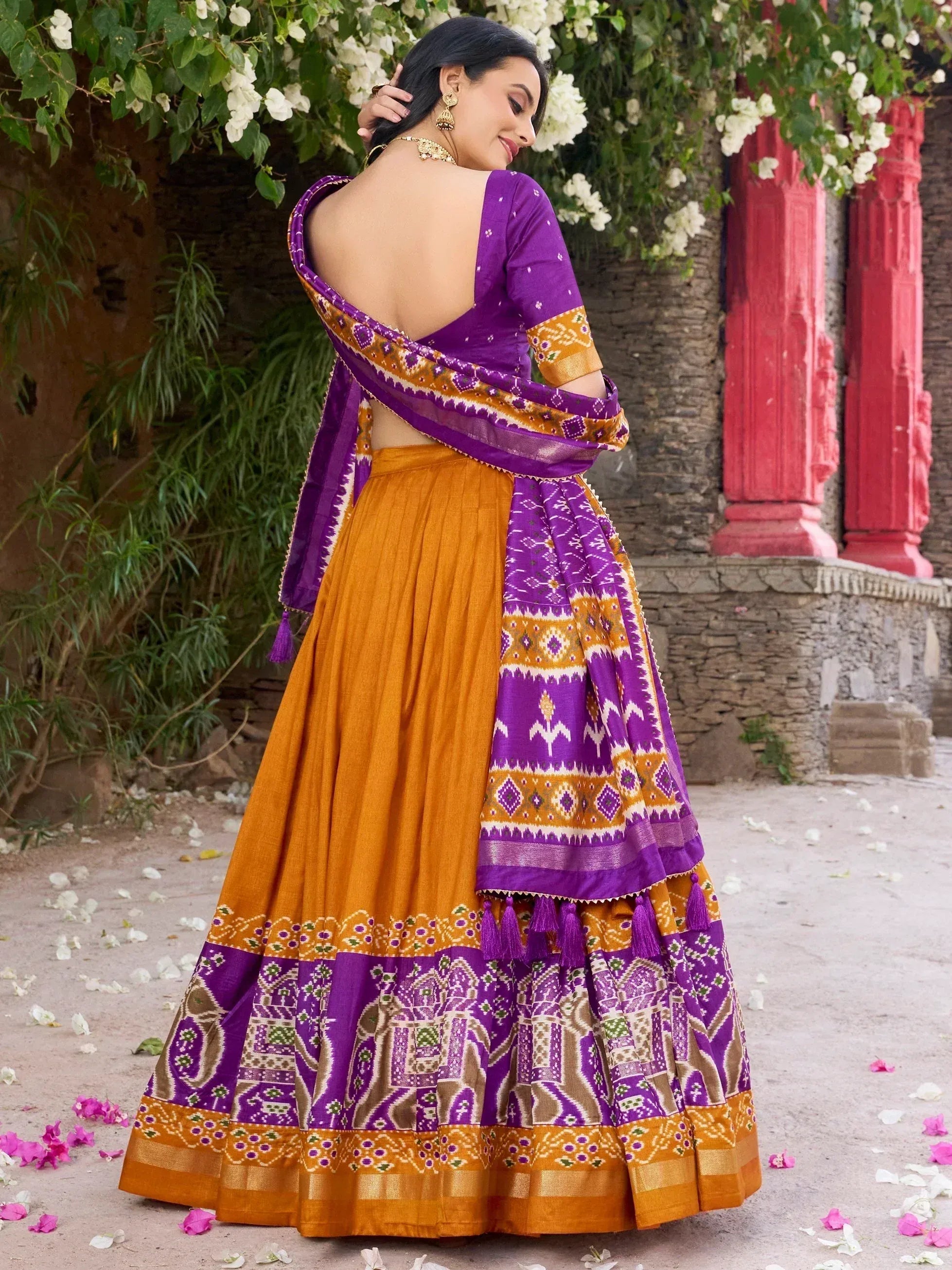 Purple and mustard Indian lehenga choli with backless blouse, ethnic print, and tassels, styled outdoors with flowers.