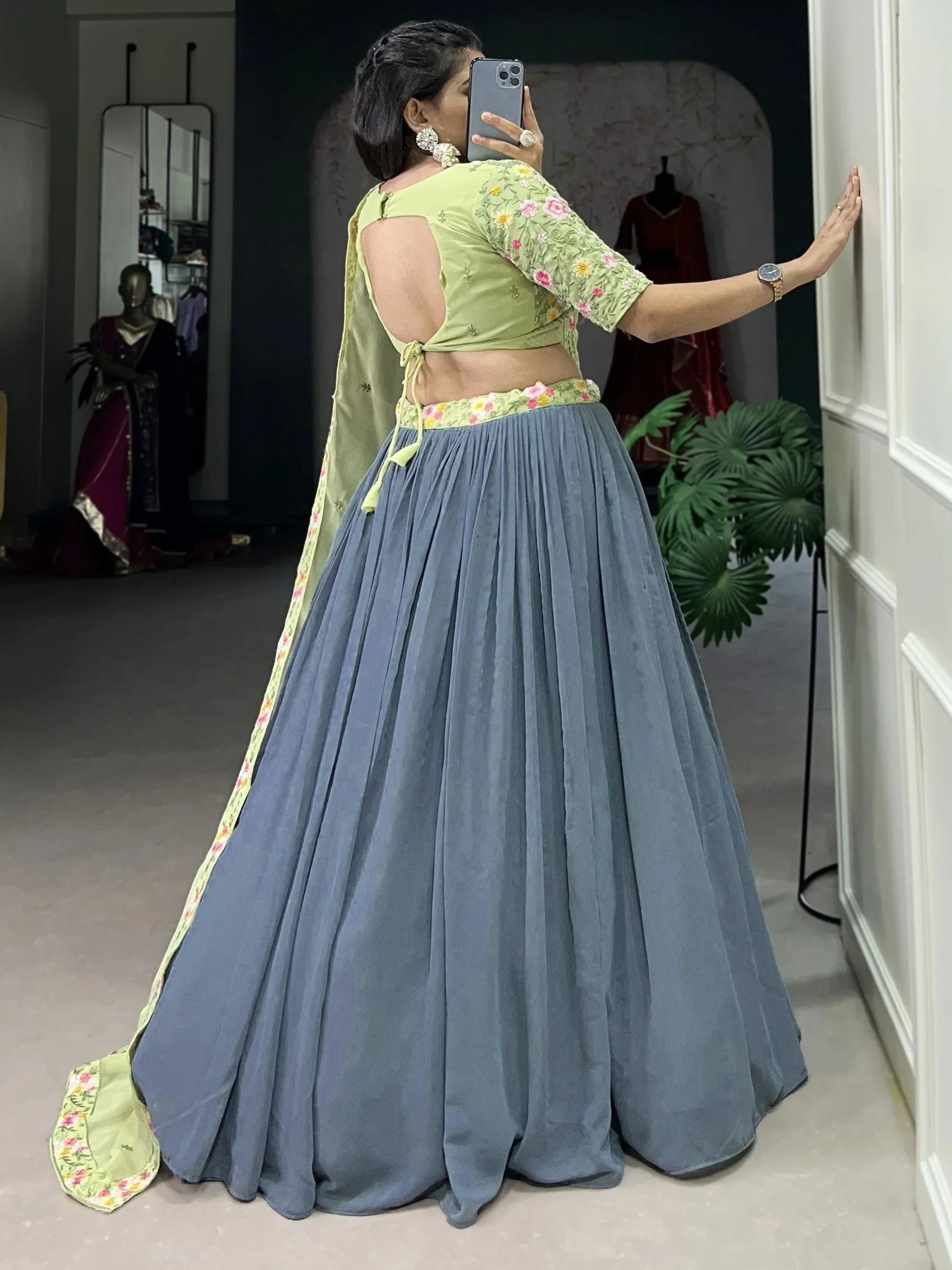 Woman in green floral blouse and blue pleated lehenga, taking mirror selfie in boutique