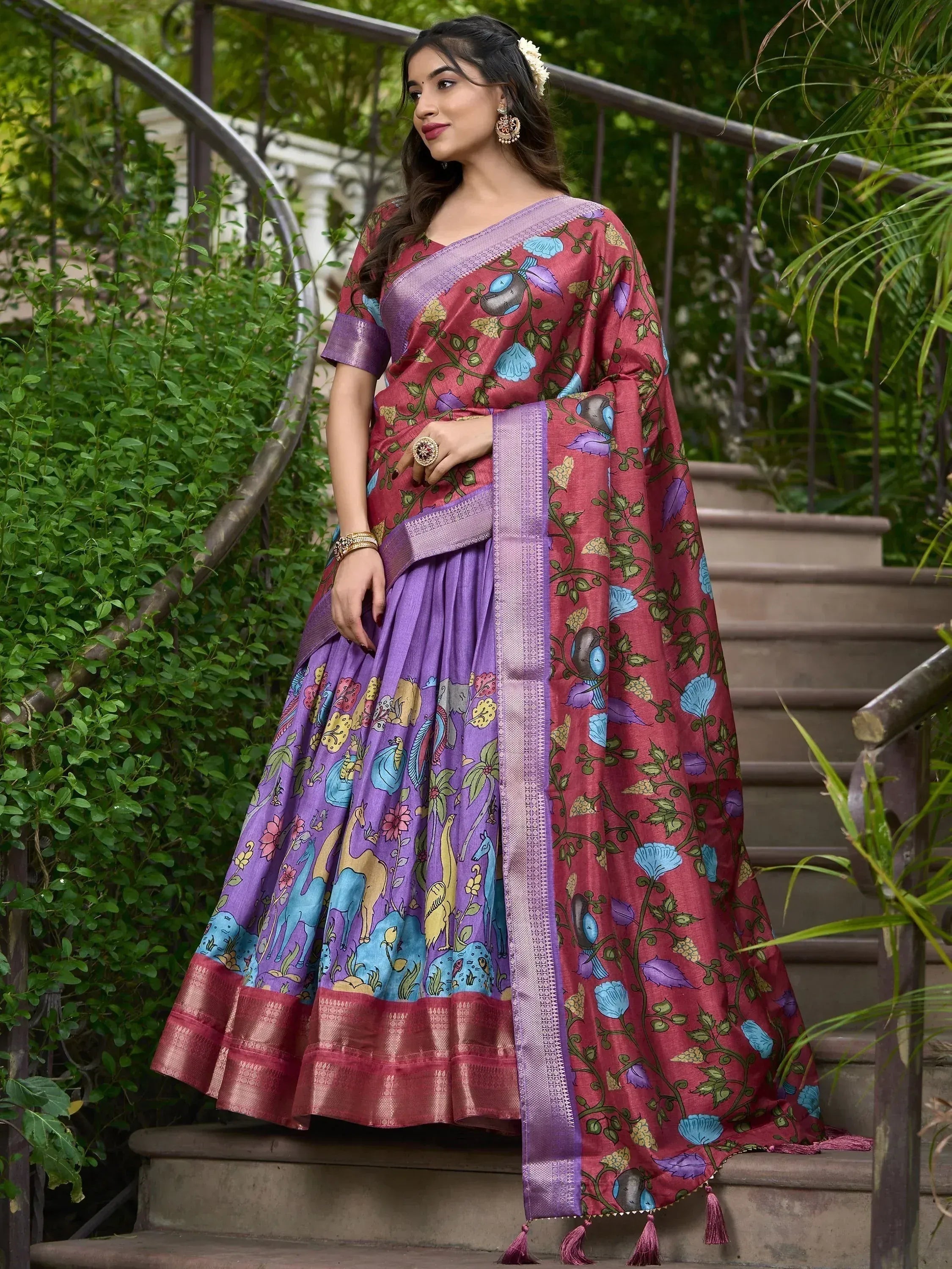 Woman in purple and maroon printed ethnic saree with floral and animal motifs, standing on garden stairs