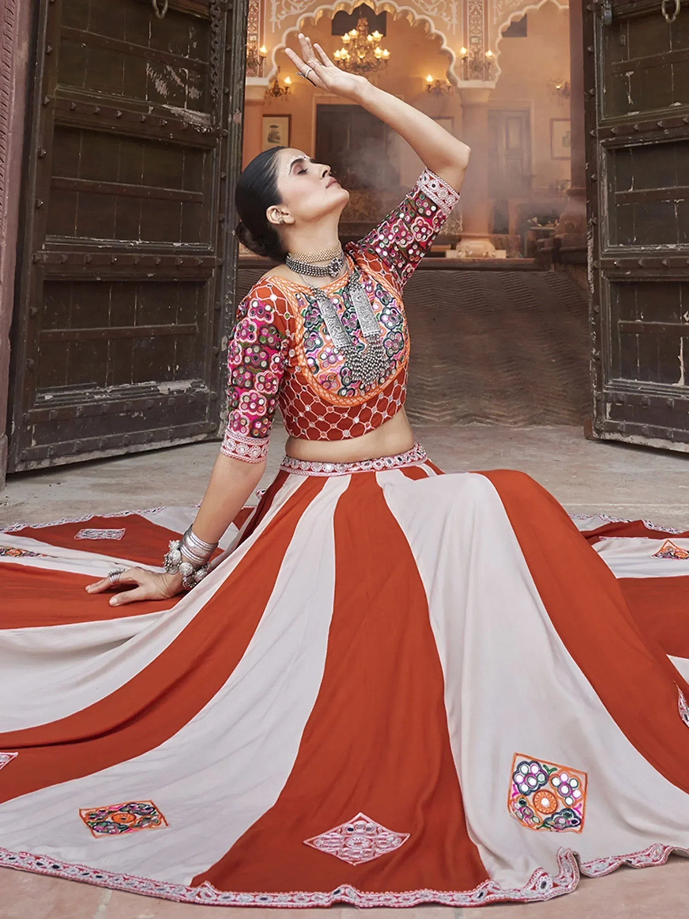 Woman in orange and white embroidered lehenga choli with mirror work, posing gracefully in traditional Indian setting