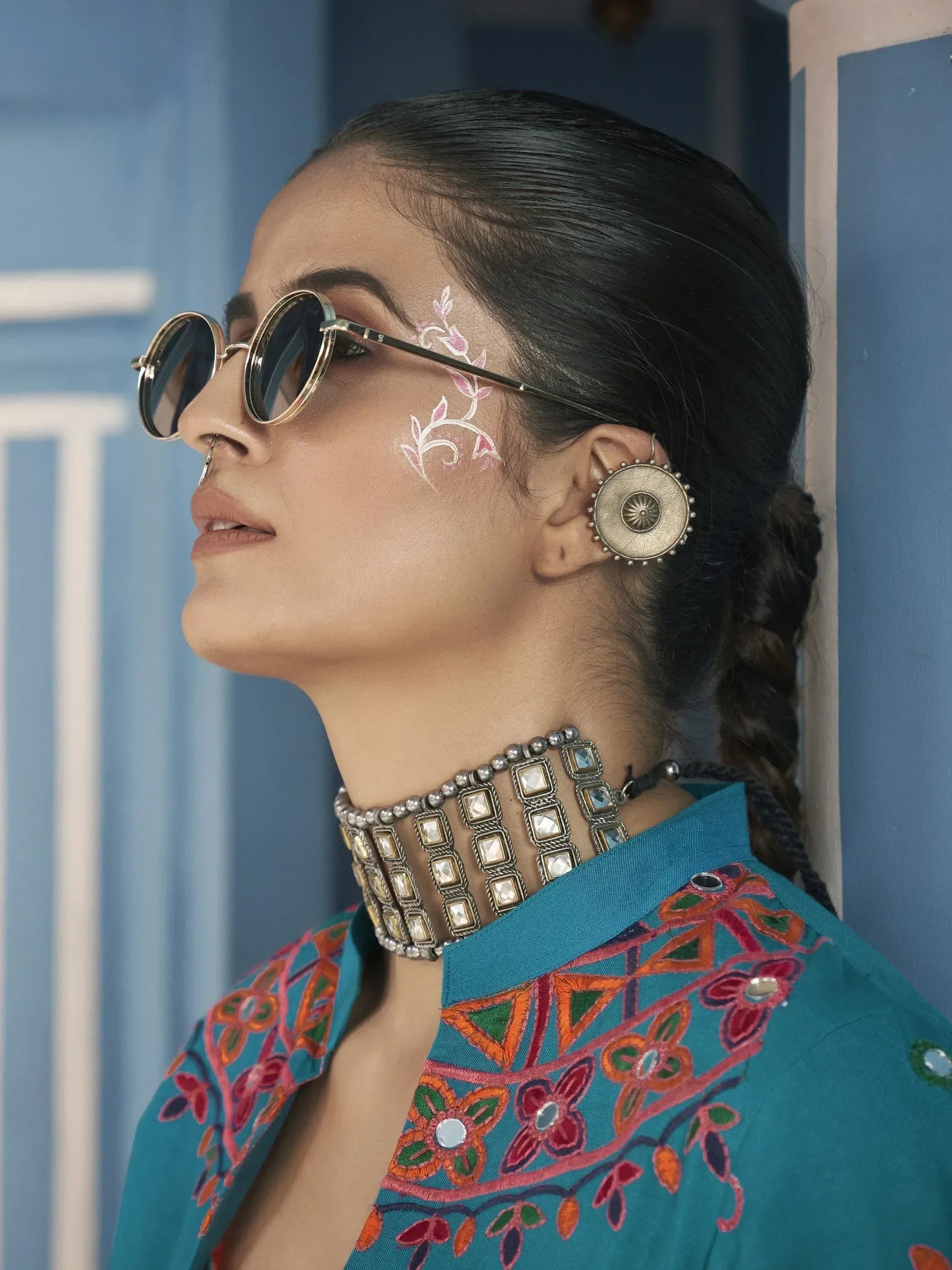 Woman in ethnic embroidered teal top with floral face art, sunglasses, and statement jewelry