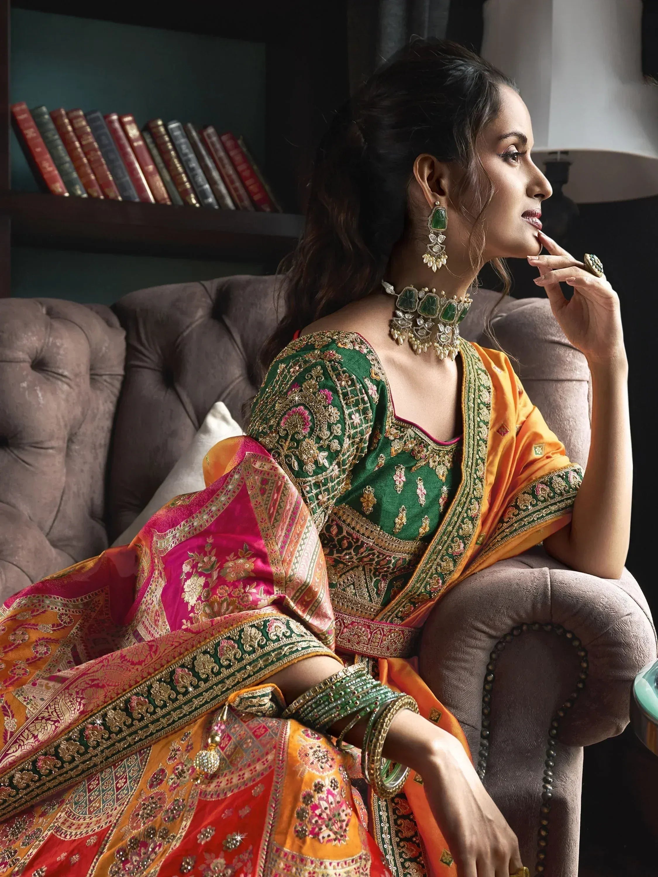 Elegant woman in green and orange embroidered lehenga choli with traditional jewelry, seated on a velvet sofa