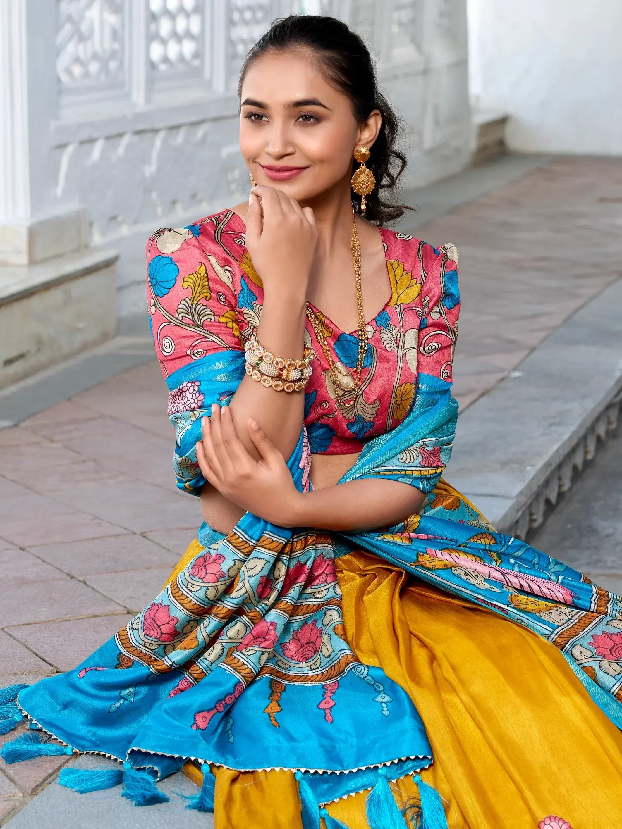 Woman in vibrant printed lehenga choli with blue dupatta, gold jewelry, sitting outdoors