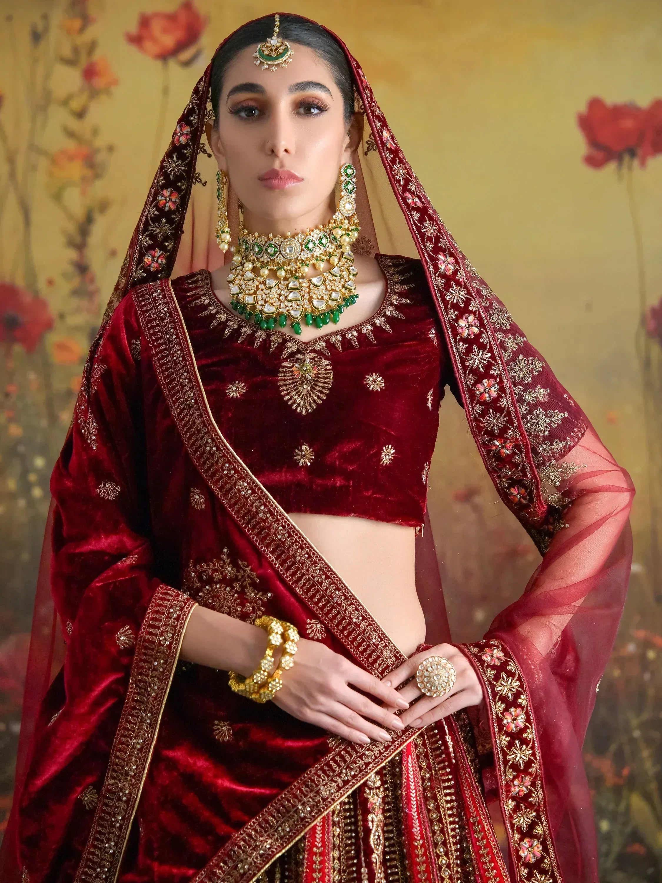 Elegant maroon velvet lehenga choli with gold embroidery and green stone jewelry for Indian weddings