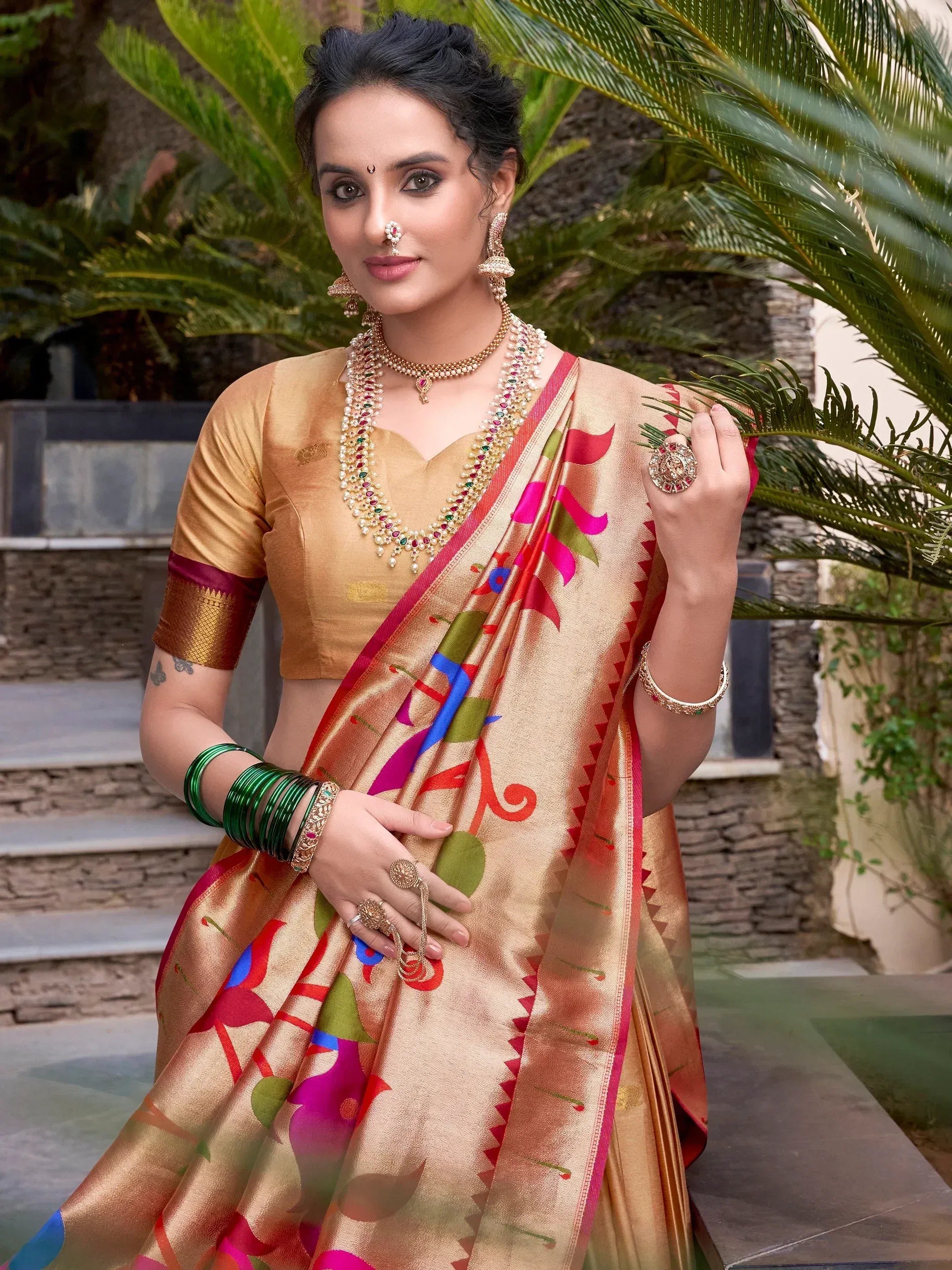 Woman in gold saree with colorful floral print, traditional Indian jewelry, and green bangles, outdoors near palm leaves.