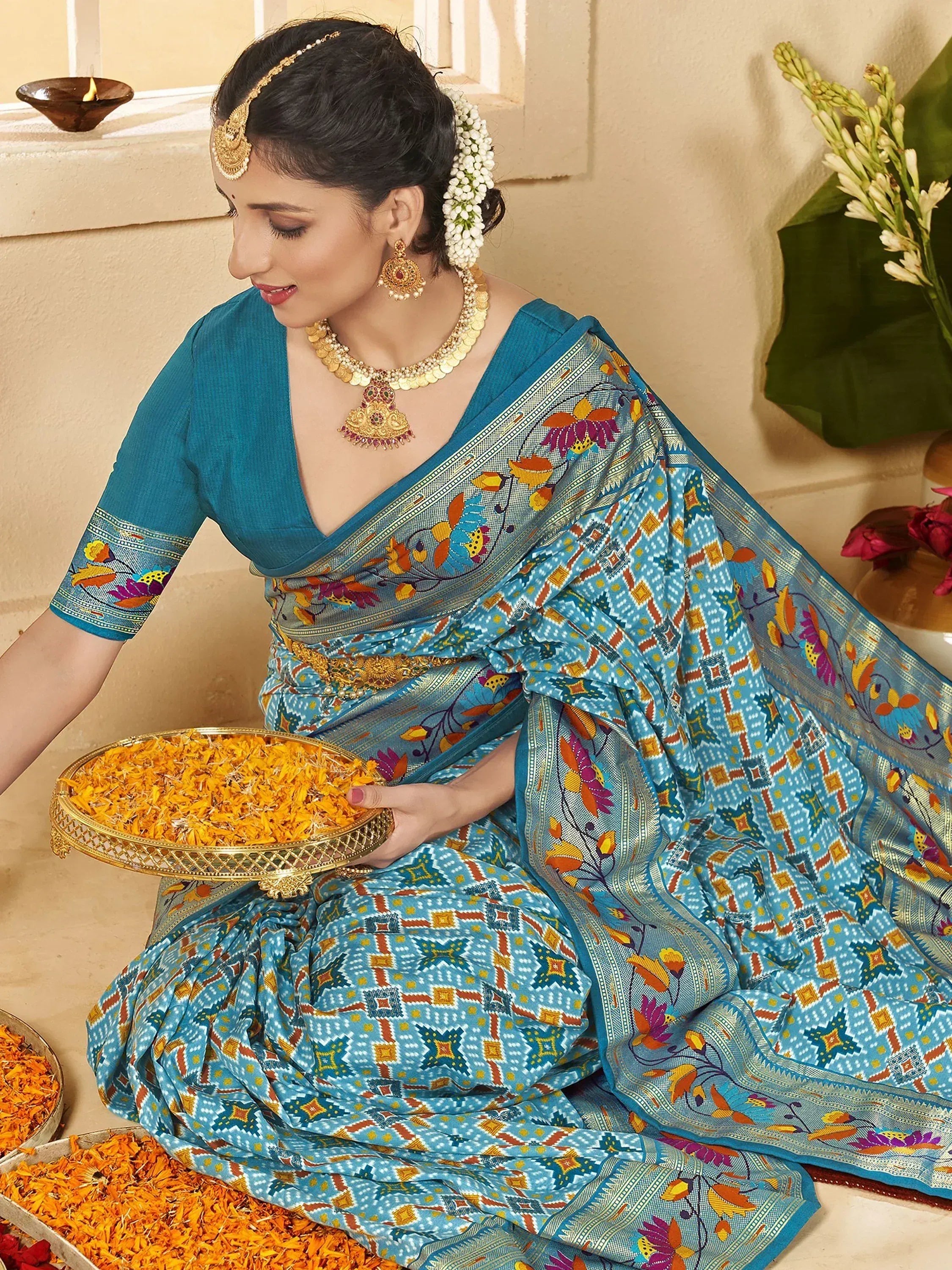 Woman in blue printed saree with floral border, gold jewelry, holding marigold petals for Indian ethnic wear