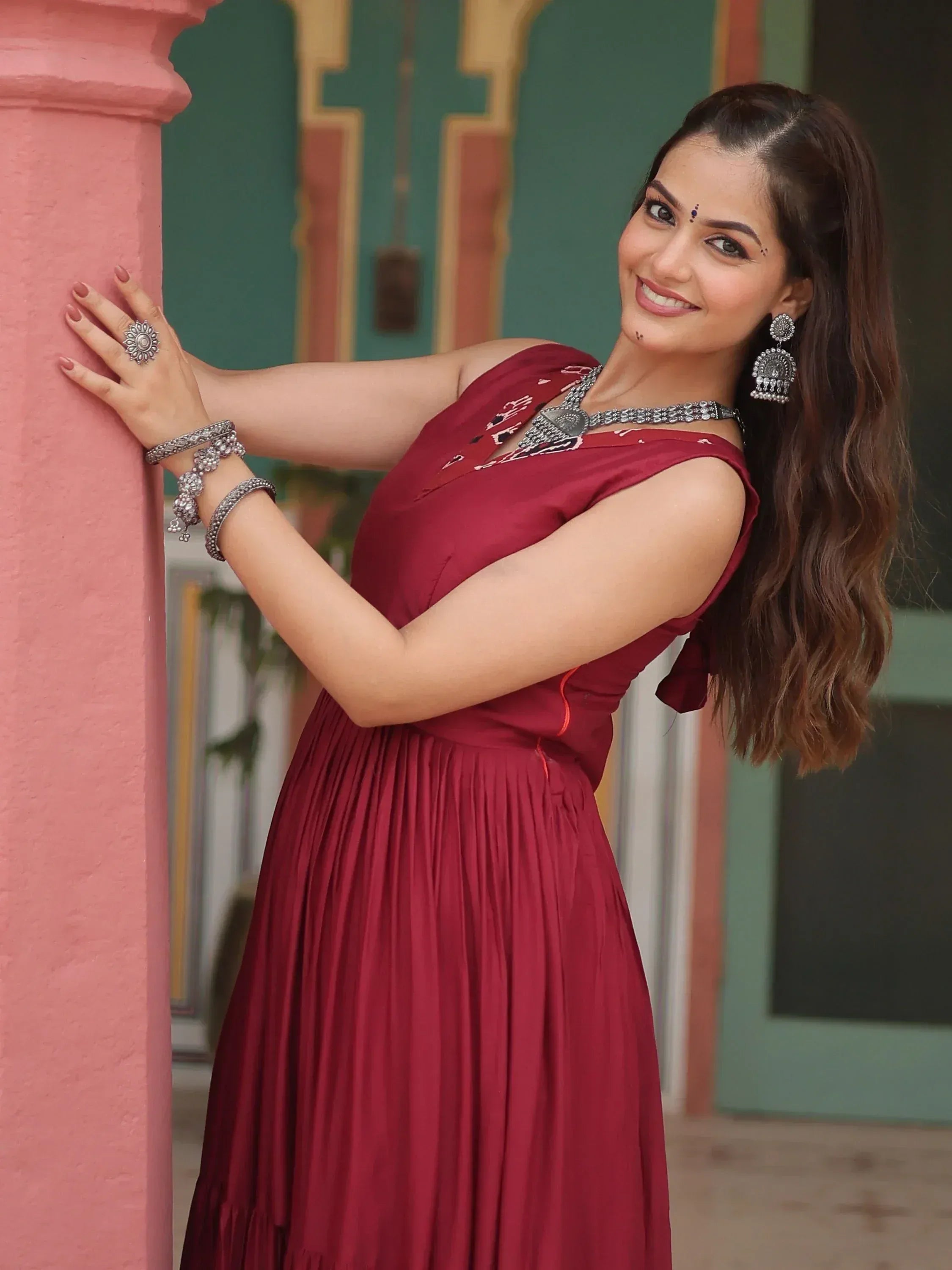 Woman in maroon ethnic gown with silver jewelry, leaning on pink pillar, Indian designer wear