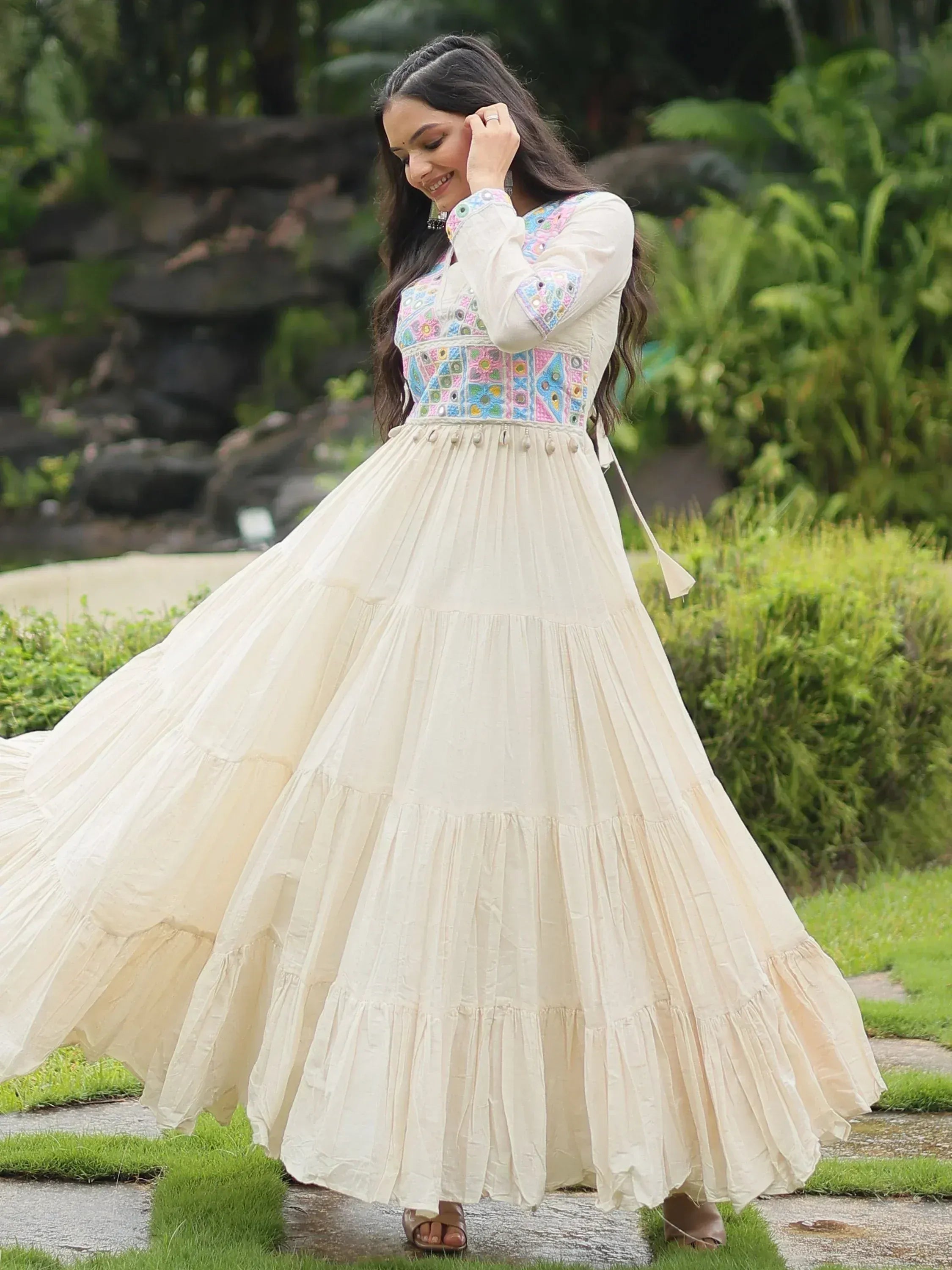 Woman in cream embroidered ethnic gown with flared skirt, posing outdoors in greenery