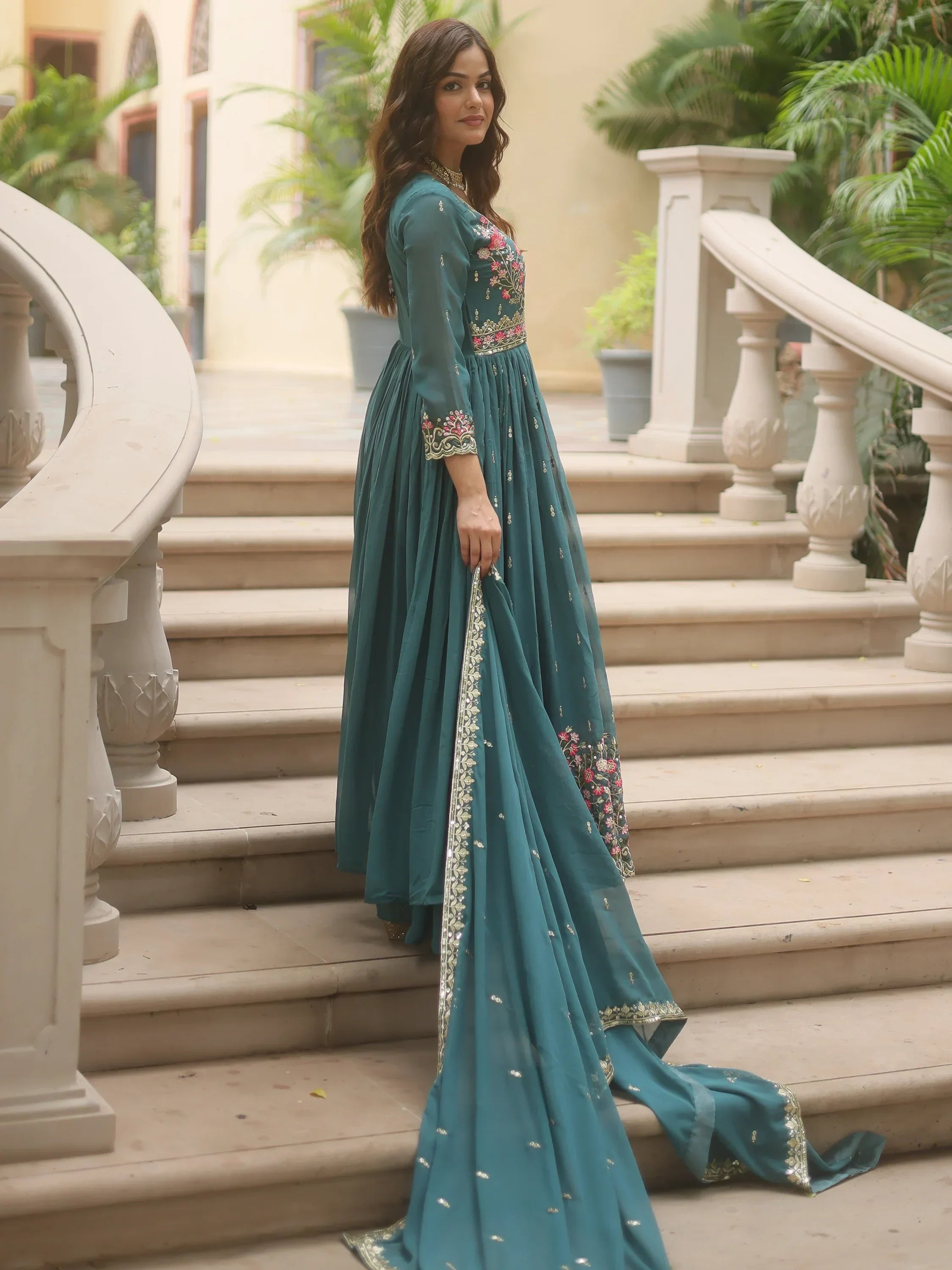 Woman wearing teal embroidered Anarkali suit with dupatta standing on outdoor stairs