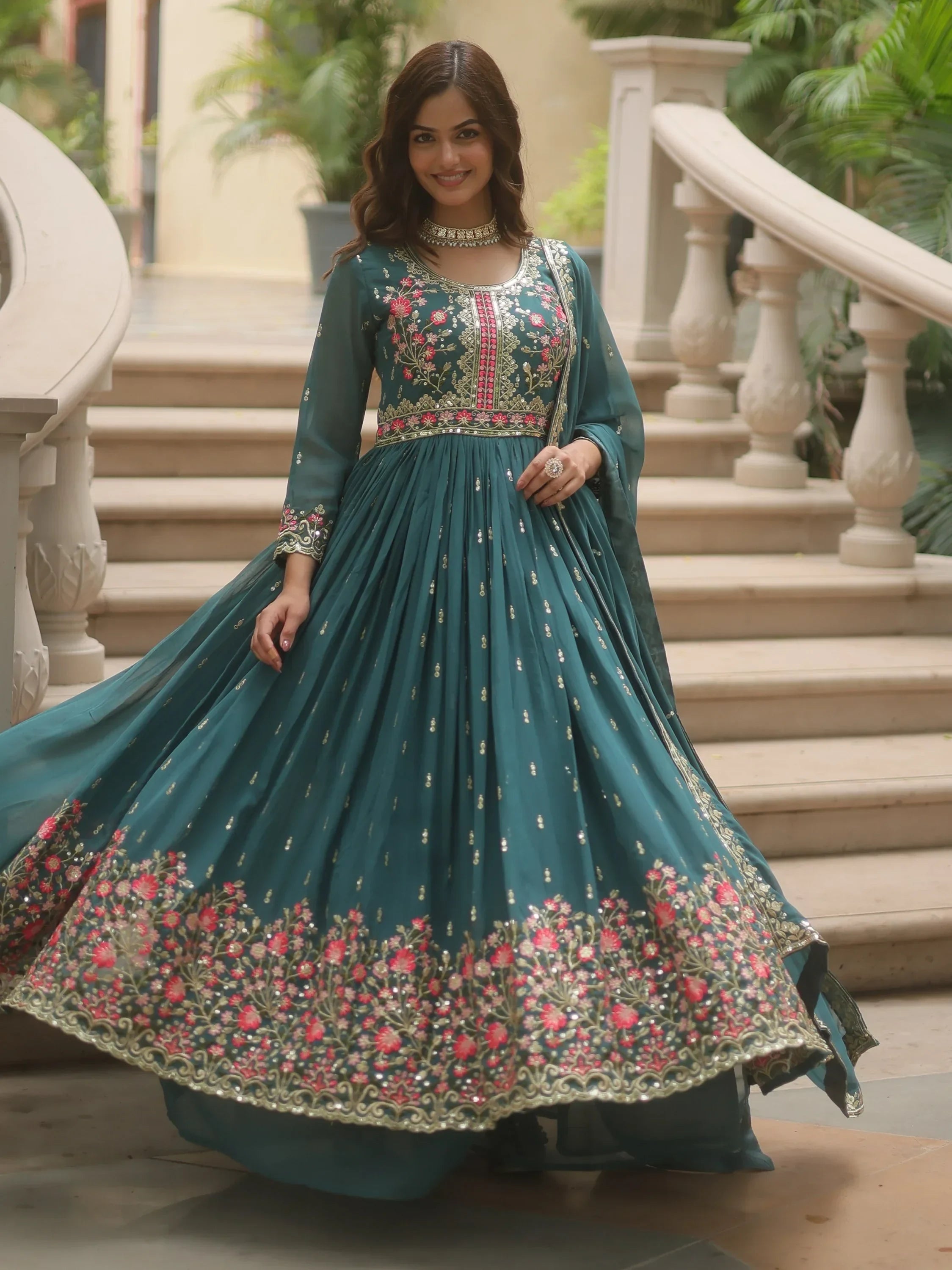 Woman in teal embroidered ethnic gown with floral details, standing on marble stairs