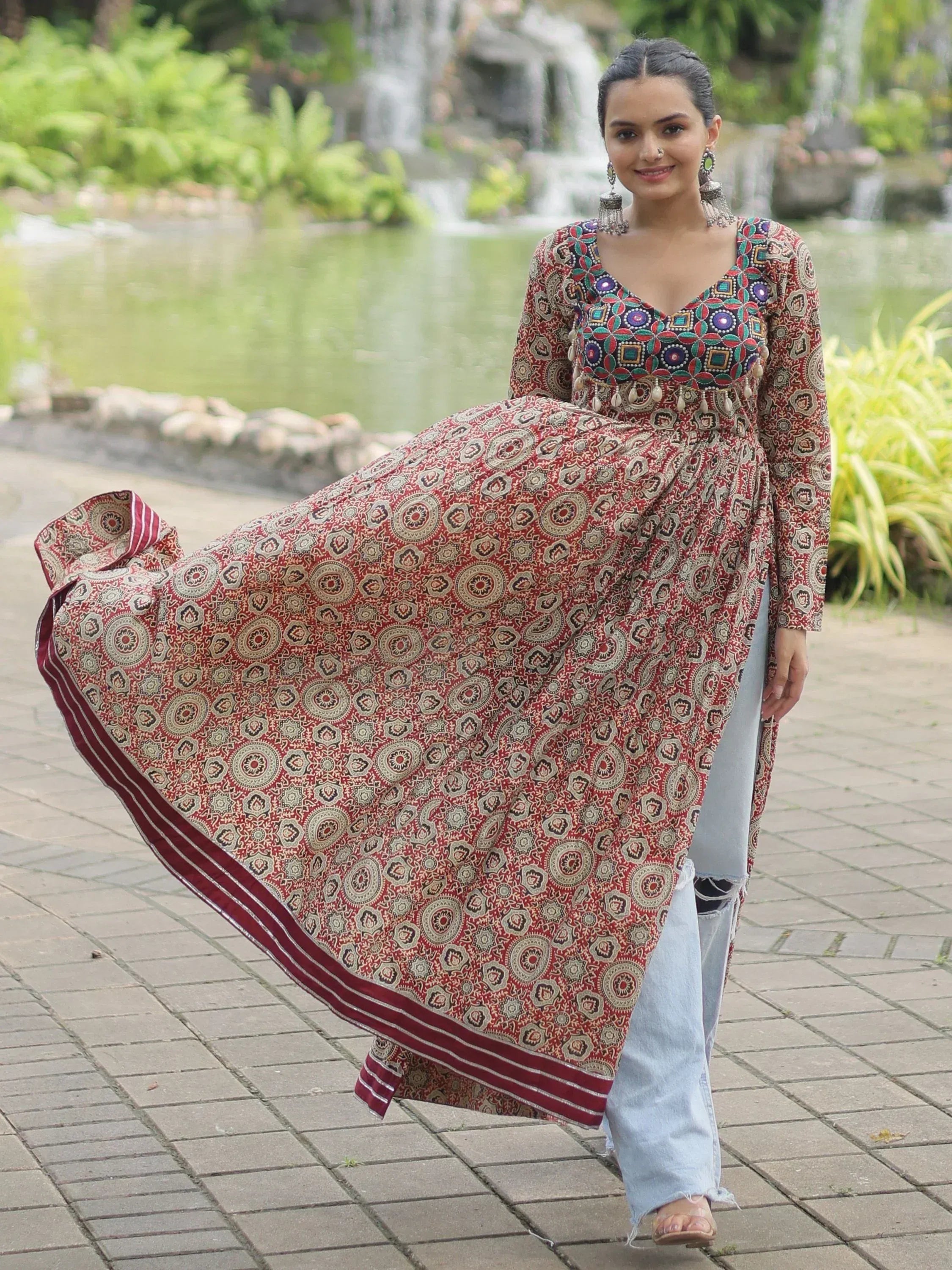 Woman in red printed ethnic gown with mirror work, paired with jeans, walking near garden pond