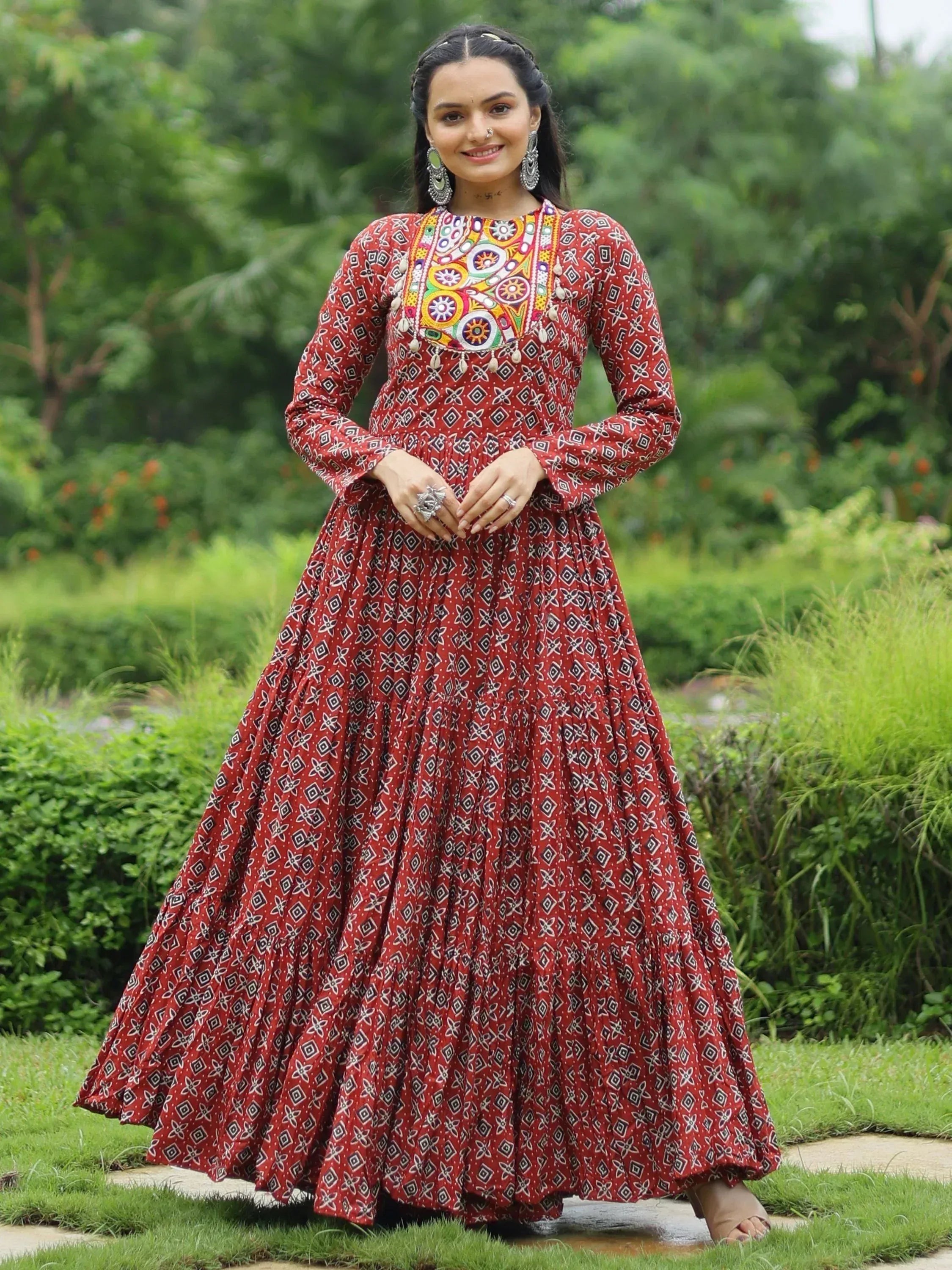 Woman in red printed ethnic gown with colorful embroidery, standing outdoors in garden