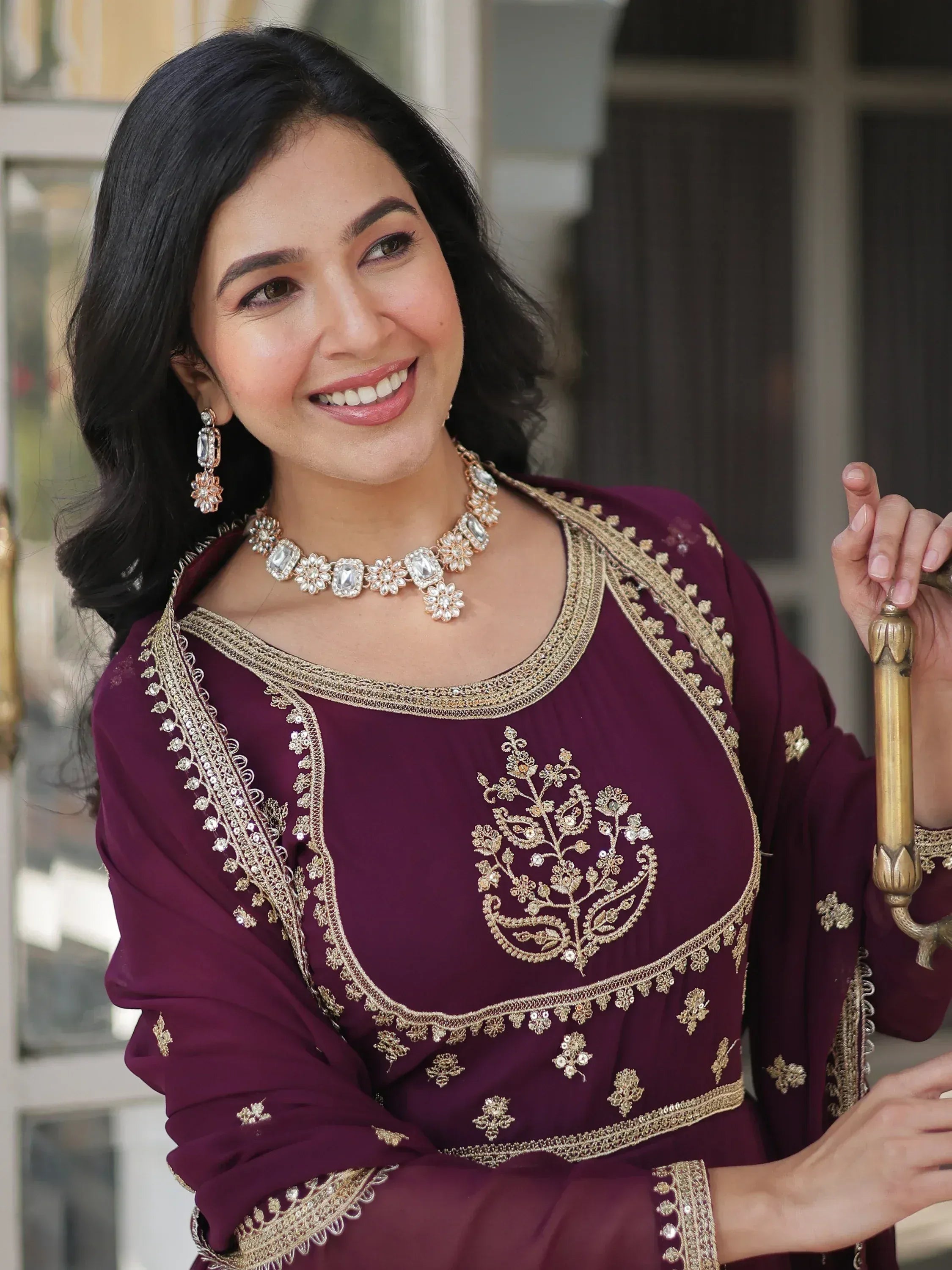 Woman in maroon ethnic gown with gold embroidery and jewelry, perfect for Indian festive occasions.