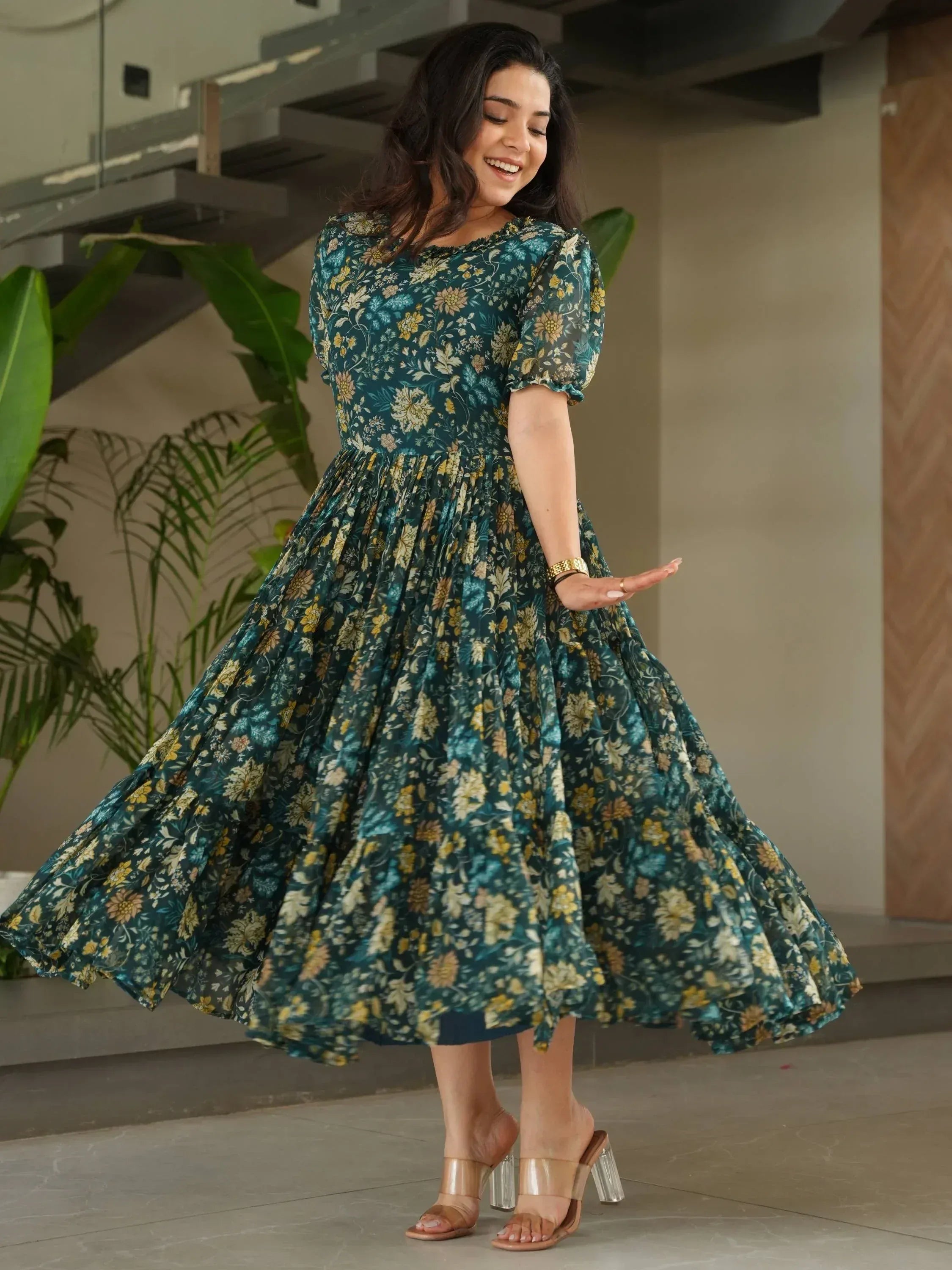 Woman in teal floral print ethnic gown with puff sleeves, twirling indoors near stairs and plants
