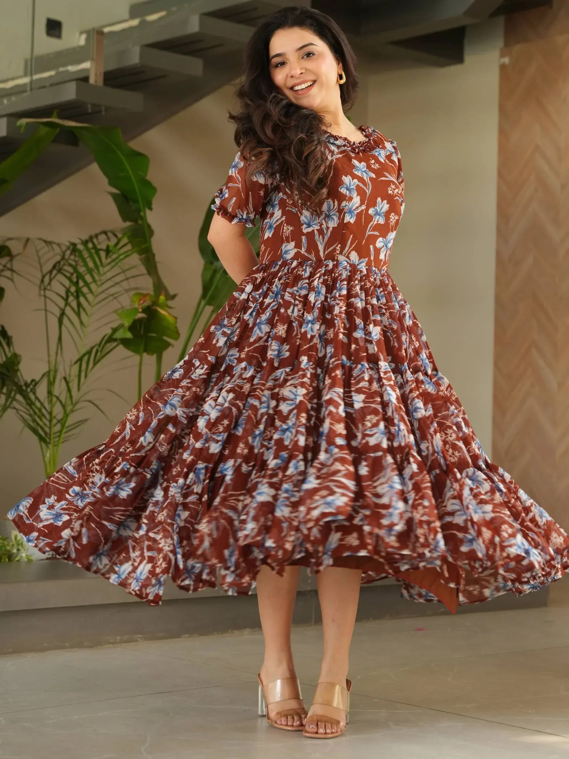 Woman in brown floral ethnic gown with puff sleeves, twirling indoors near stairs and plants