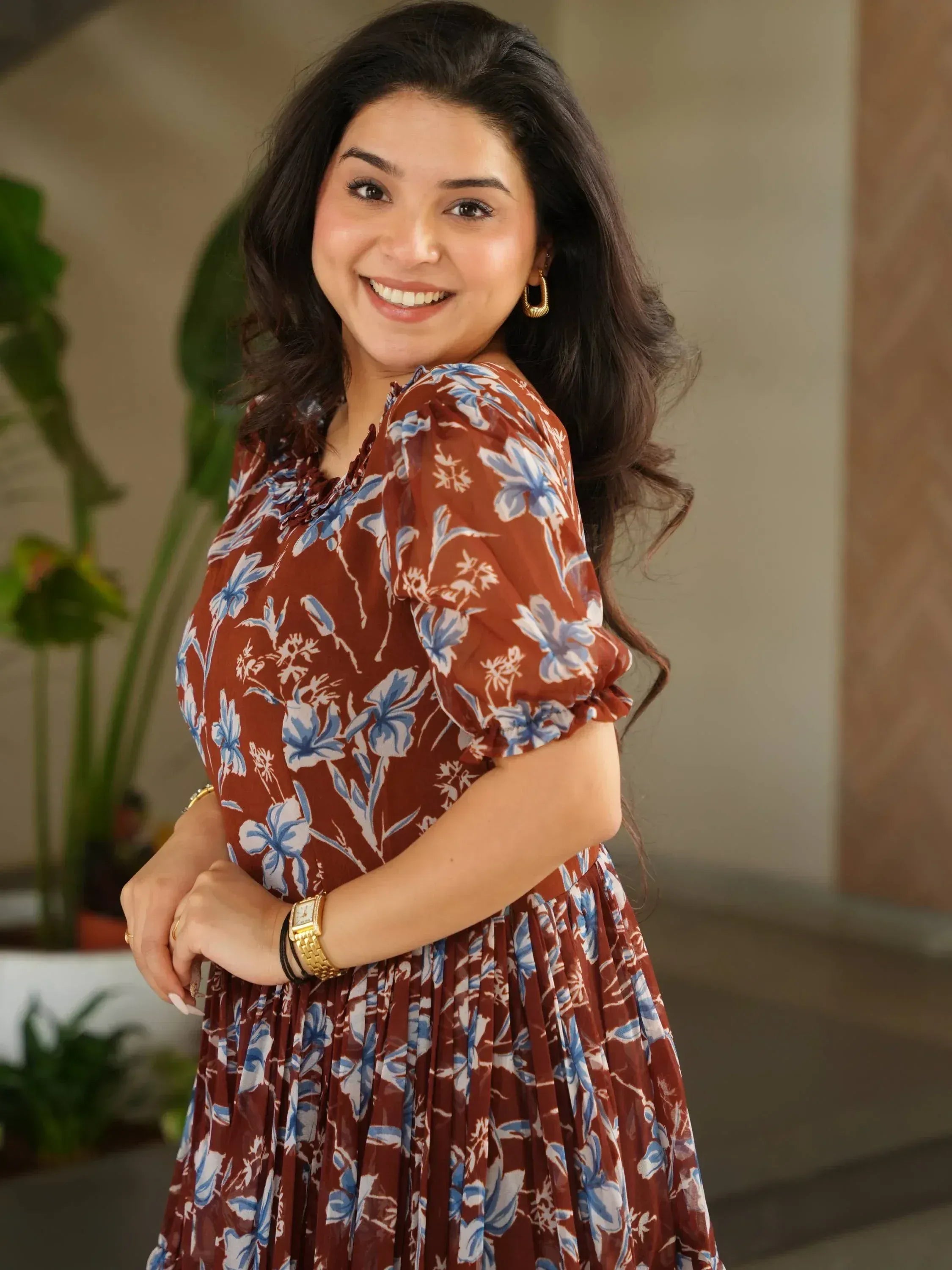 Woman in brown floral ethnic gown with blue prints, gold accessories, smiling indoors