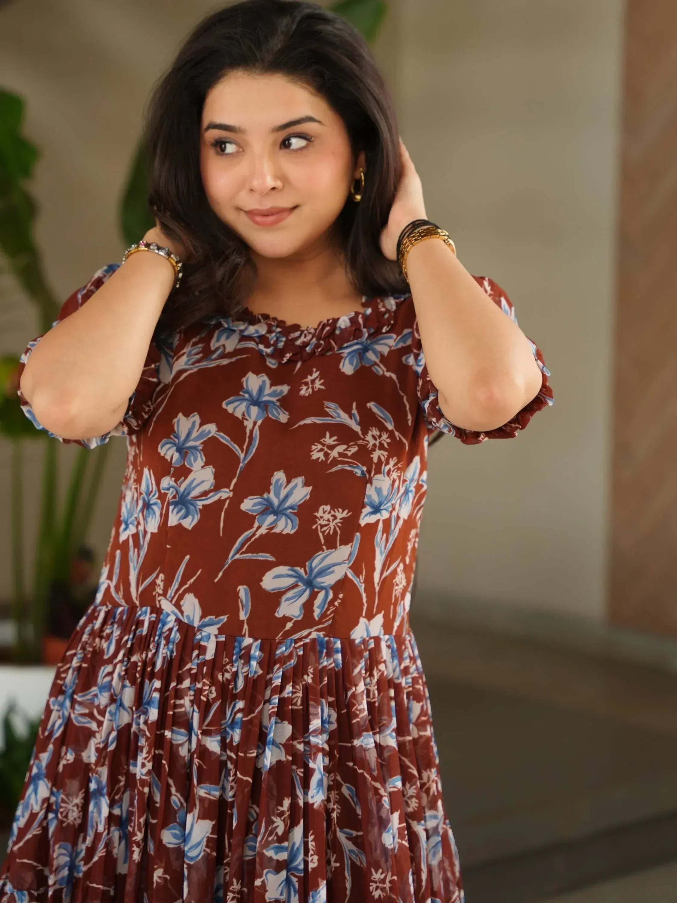 Woman in brown floral ethnic gown with blue print, ruffled sleeves, and pleated skirt, styled with gold jewelry.