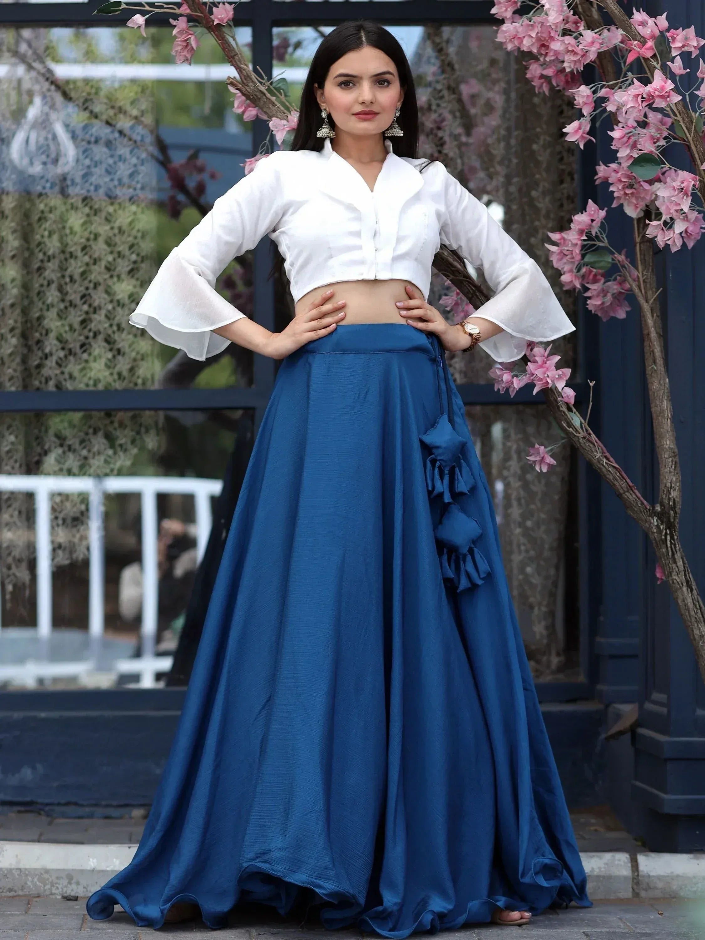 Woman in white crop top with bell sleeves and blue lehenga skirt, floral backdrop, ethnic wear
