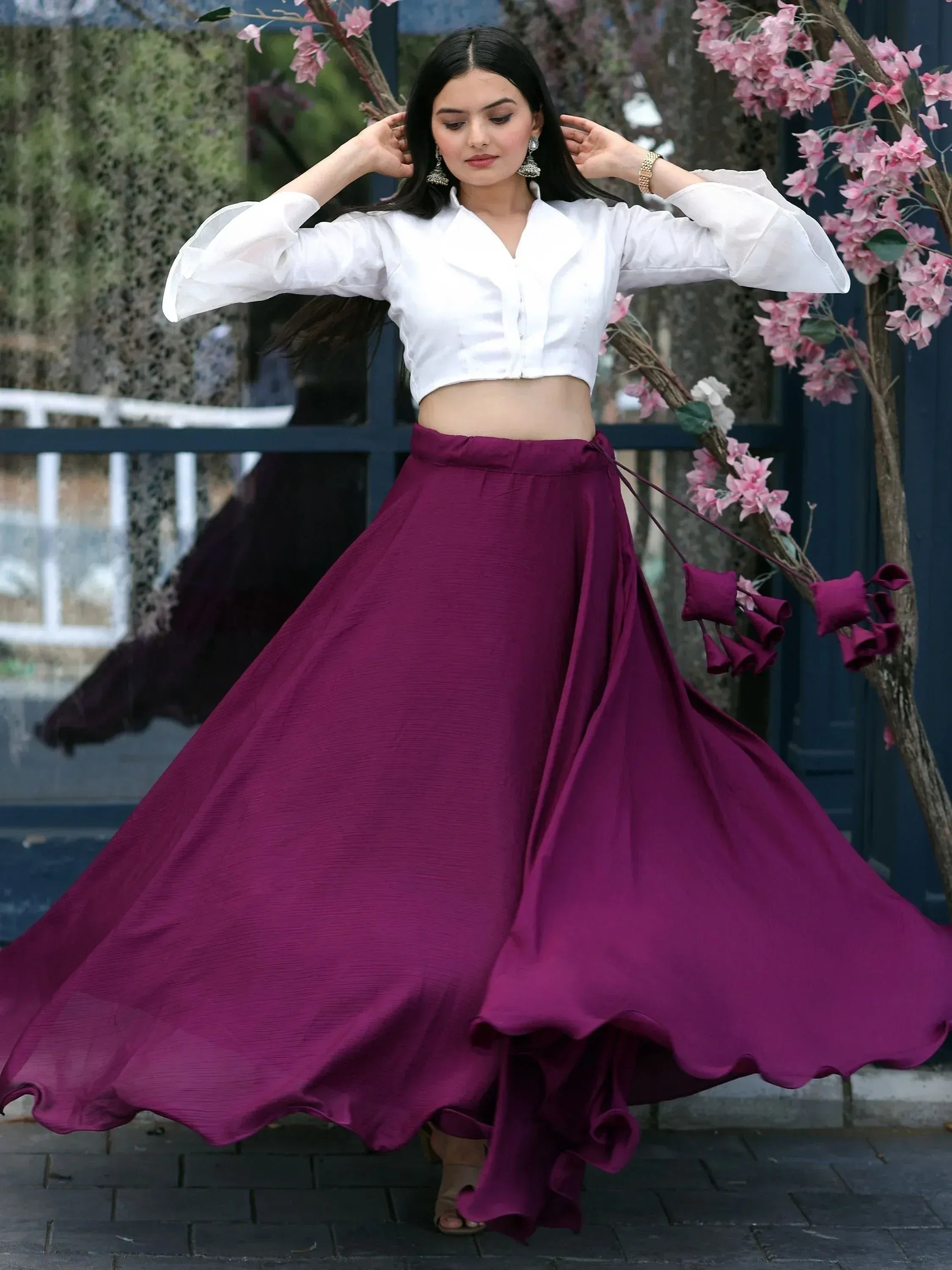 Woman in white crop top and flowing purple skirt, twirling outdoors near pink blossoms