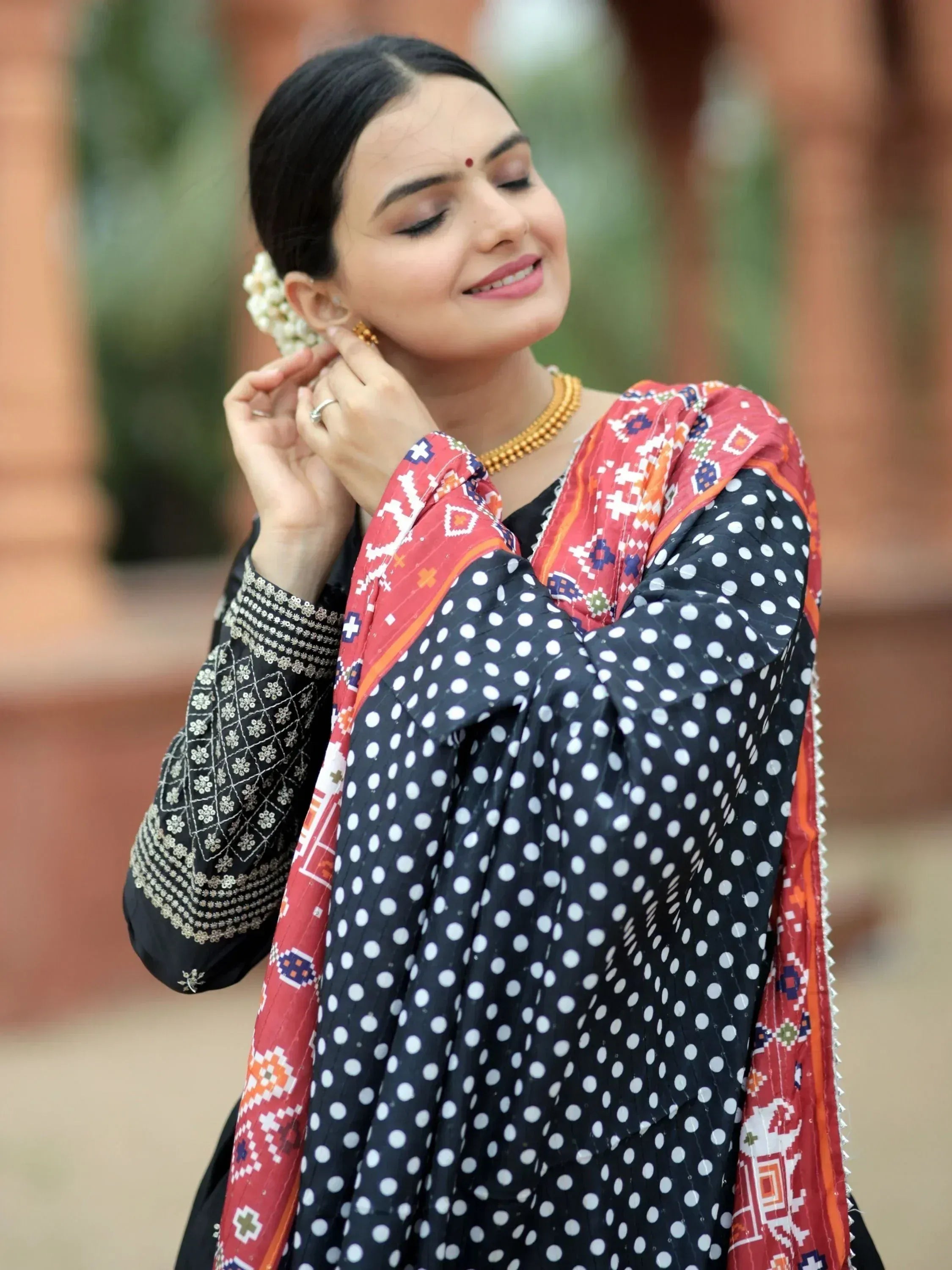 Woman in black polka dot salwar kameez with embroidered sleeves and colorful dupatta, accessorized with gold jewelry