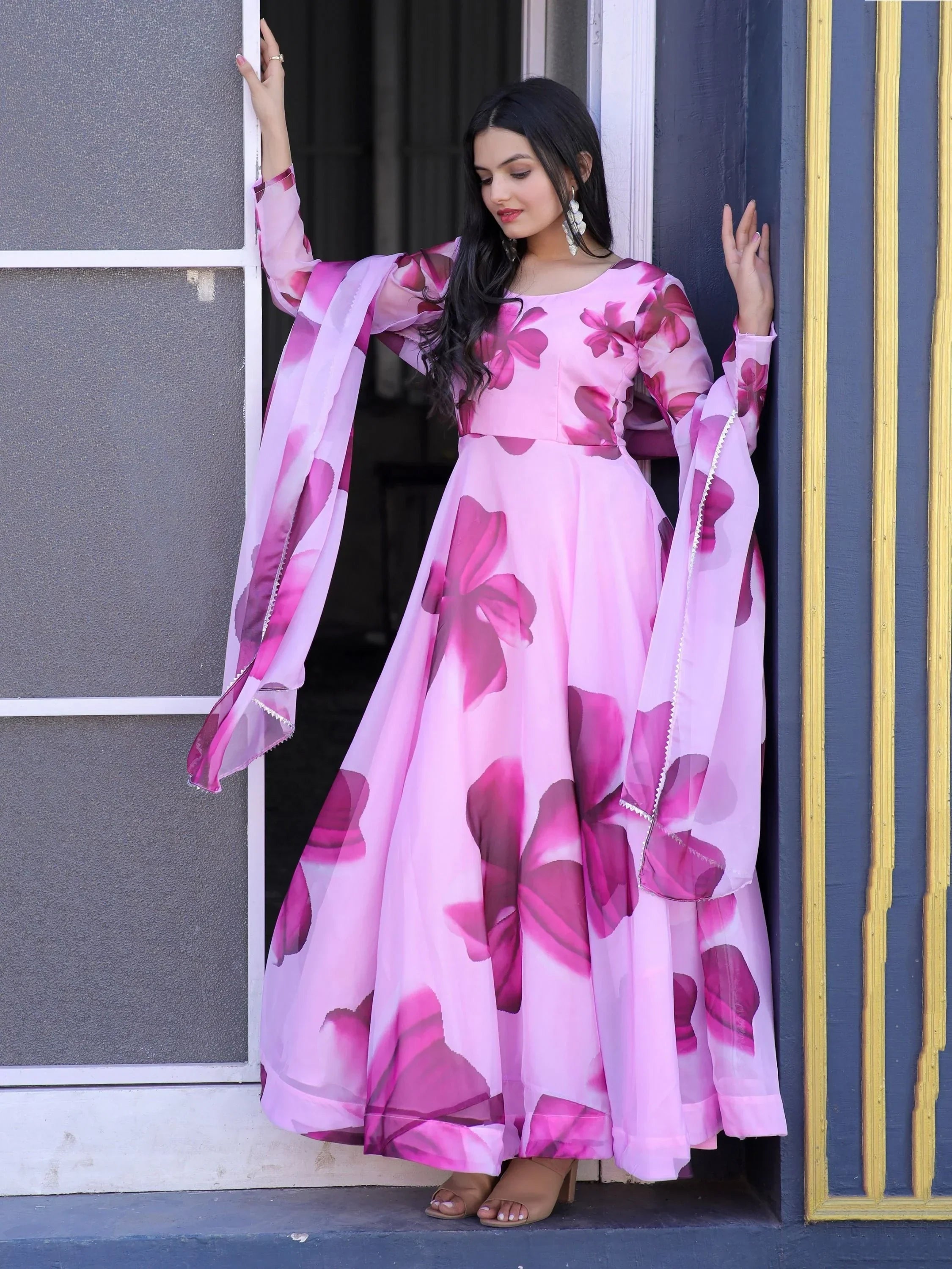 Woman wearing pink floral printed ethnic gown with dupatta, standing by a doorway