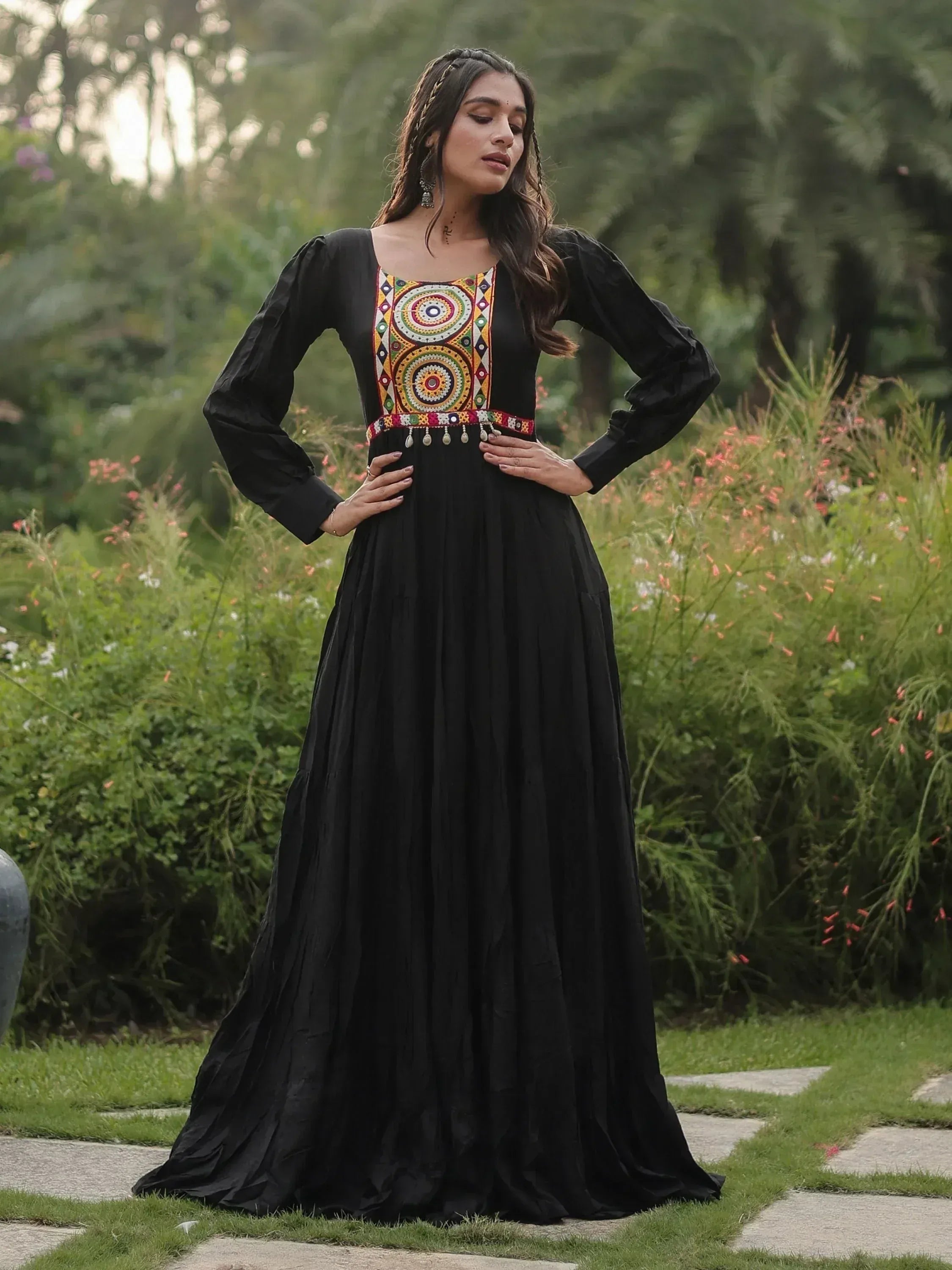 Black ethnic gown with colorful embroidery, long sleeves, and pleated skirt, styled in a garden setting.