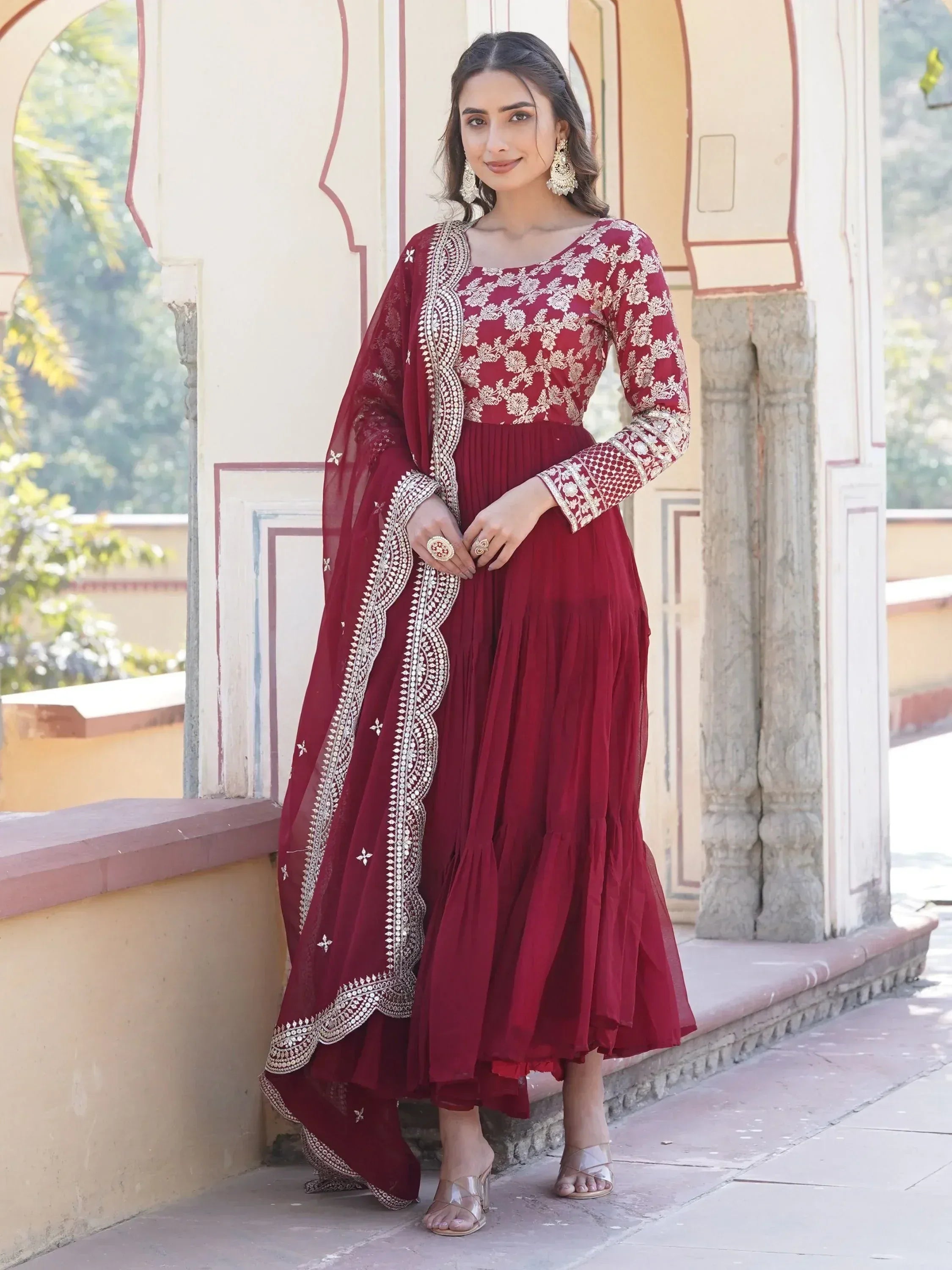 Woman in maroon ethnic gown with gold embroidery and dupatta, standing outdoors