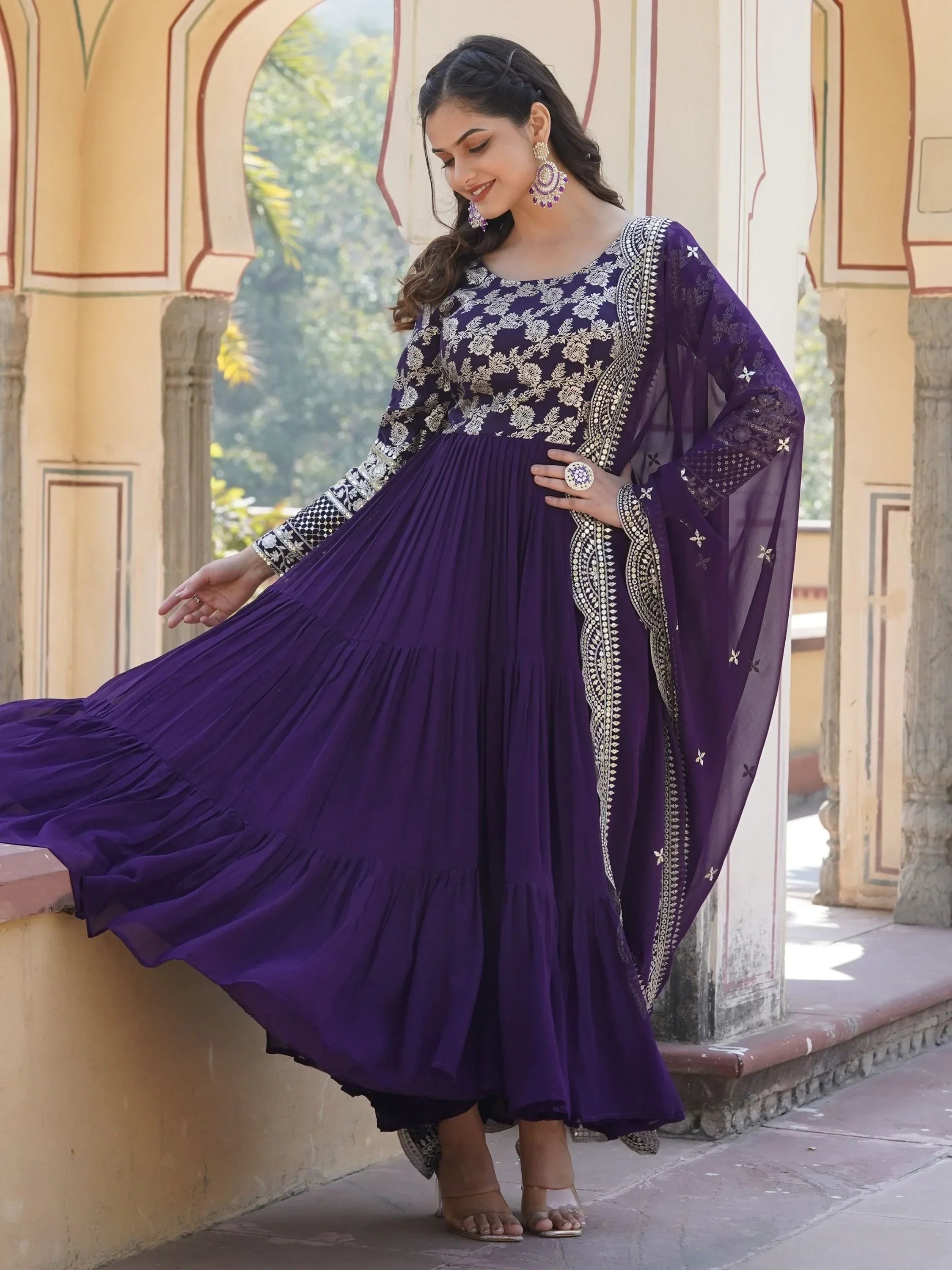 Woman in a purple embroidered Anarkali gown with dupatta, standing in a traditional outdoor setting.